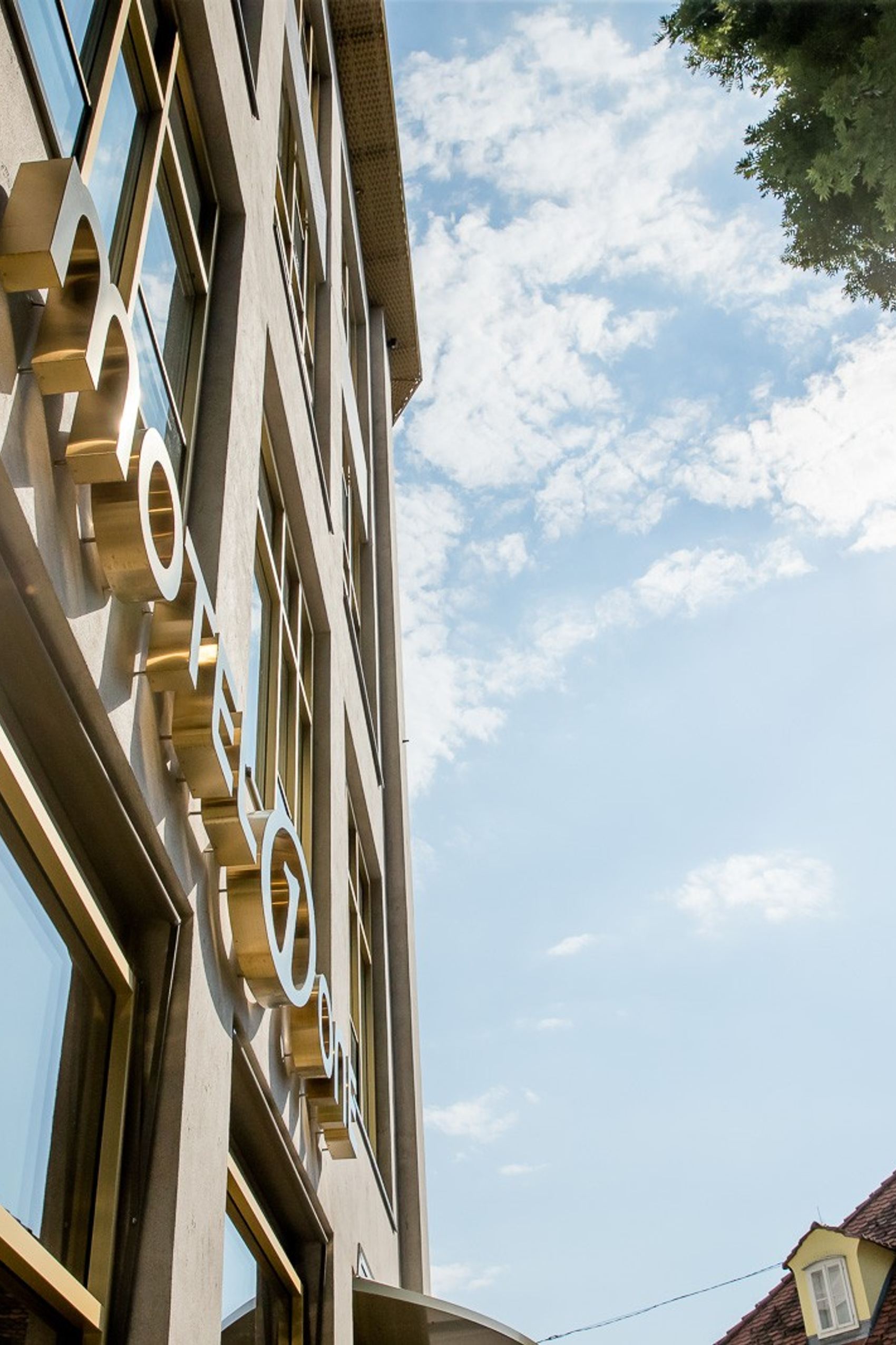 Blick nach oben auf die moderne Fassade eines Hotels mit großem, reflektierendem Fenster, darunter die Hotelbeschriftung. Im Hintergrund sind Dachkuppeln traditioneller Häuser und ein sonniger Himmel mit wenigen Wolken zu sehen.