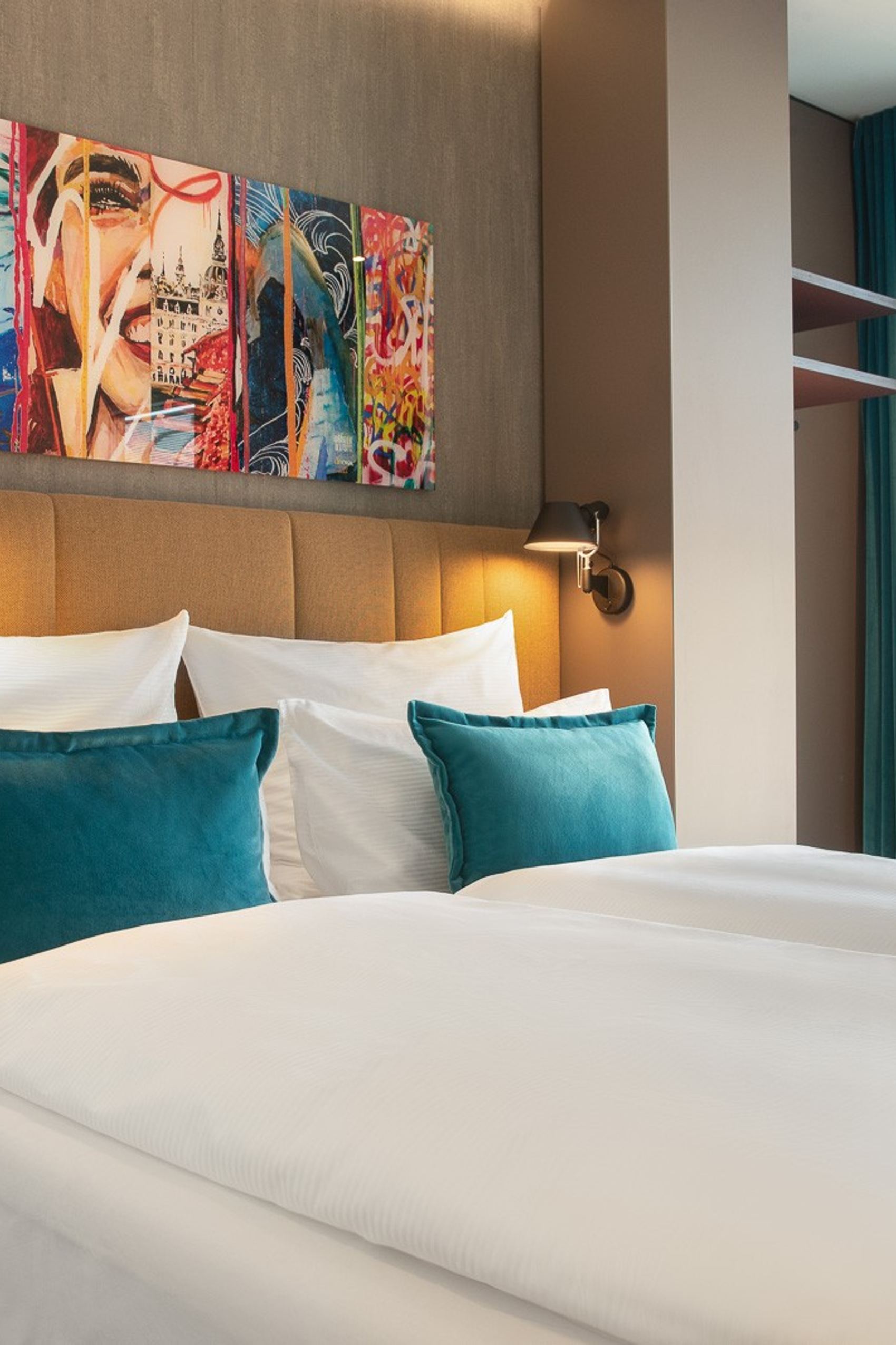 Hotelzimmer mit großem Doppelbett, weißen und türkisfarbenen Kissen, stimmungsvoller Beleuchtung und farbenfroher Kunst über dem Kopfteil. Helles Zimmer mit großem Fenster und Blick auf die Stadt.