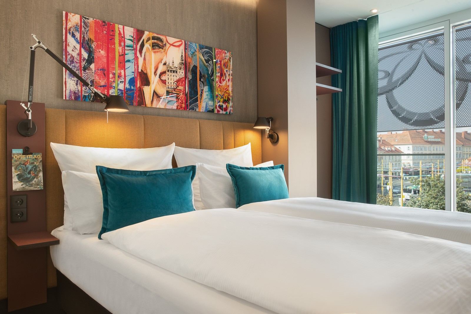 Modern eingerichtetes Hotelzimmer mit großem Doppelbett, weißen und türkisfarbenen Kissen, stimmungsvoller Beleuchtung und farbenfroher Kunst über dem Kopfteil. Helles Zimmer mit großem Fenster und Blick auf die Stadt. Modern eingerichtetes Hotelzimmer mit großem Doppelbett, weißen und türkisfarbenen Kissen, stimmungsvoller Beleuchtung und farbenfroher Kunst über dem Kopfteil. Helles Zimmer mit großem Fenster und Blick auf die Stadt.