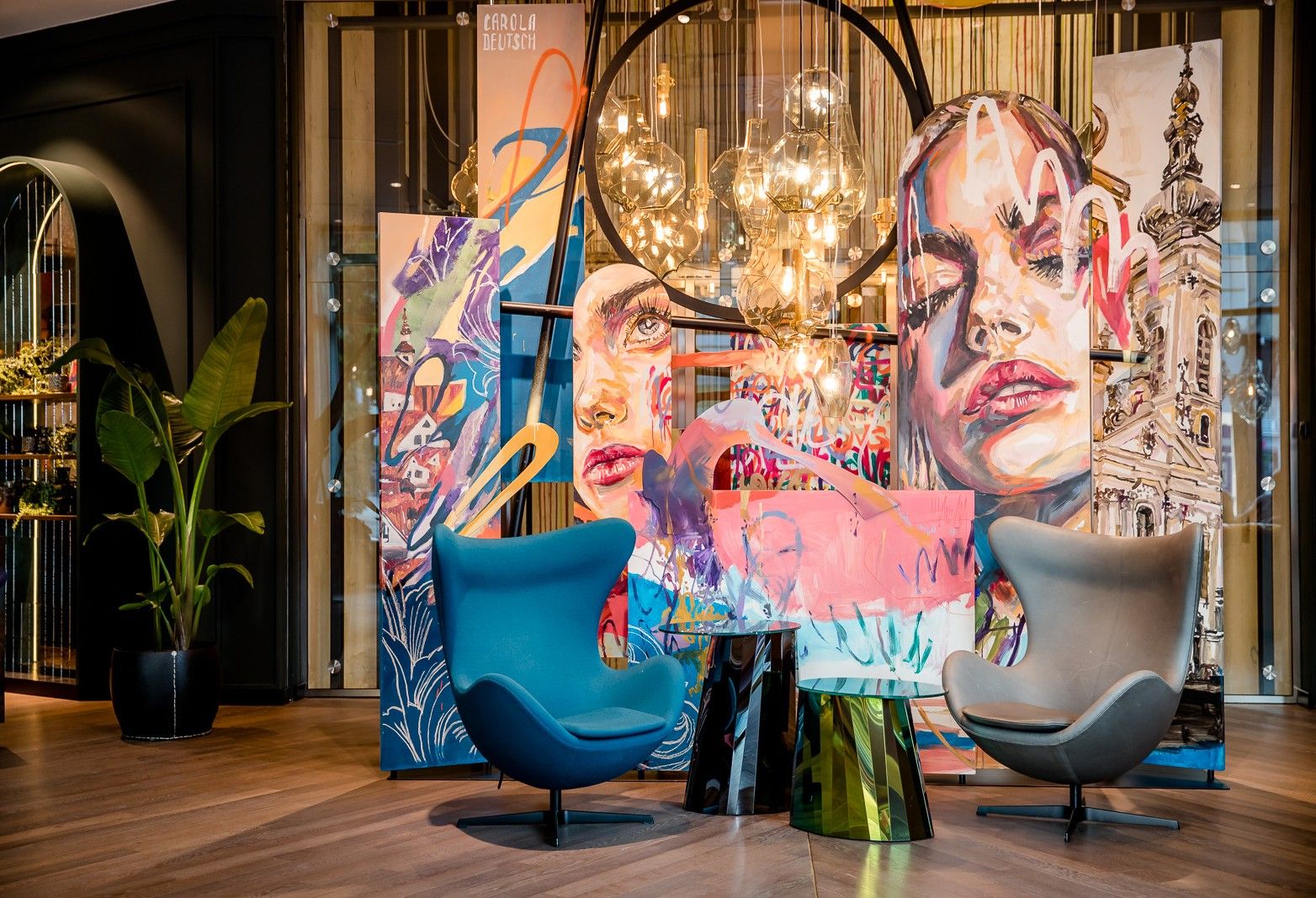 Zwei moderne, farblich unterschiedliche Sessel vor einer großen, farbenfrohen Wandkunst mit Porträts und abstrakten Elementen, beleuchtet von warmen Hängelampen, schaffen eine einladende Lounge-Atmosphäre in einem stilvollen, gemütlichen Innenraum.