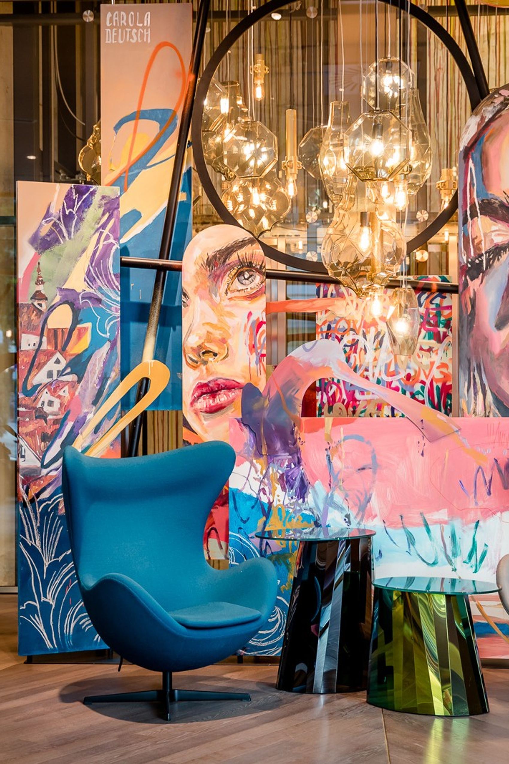 Zwei moderne, farblich unterschiedliche Sessel vor einer großen, farbenfrohen Wandkunst mit Porträts und abstrakten Elementen, beleuchtet von warmen Hängelampen, schaffen eine einladende Lounge-Atmosphäre in einem stilvollen, gemütlichen Innenraum.