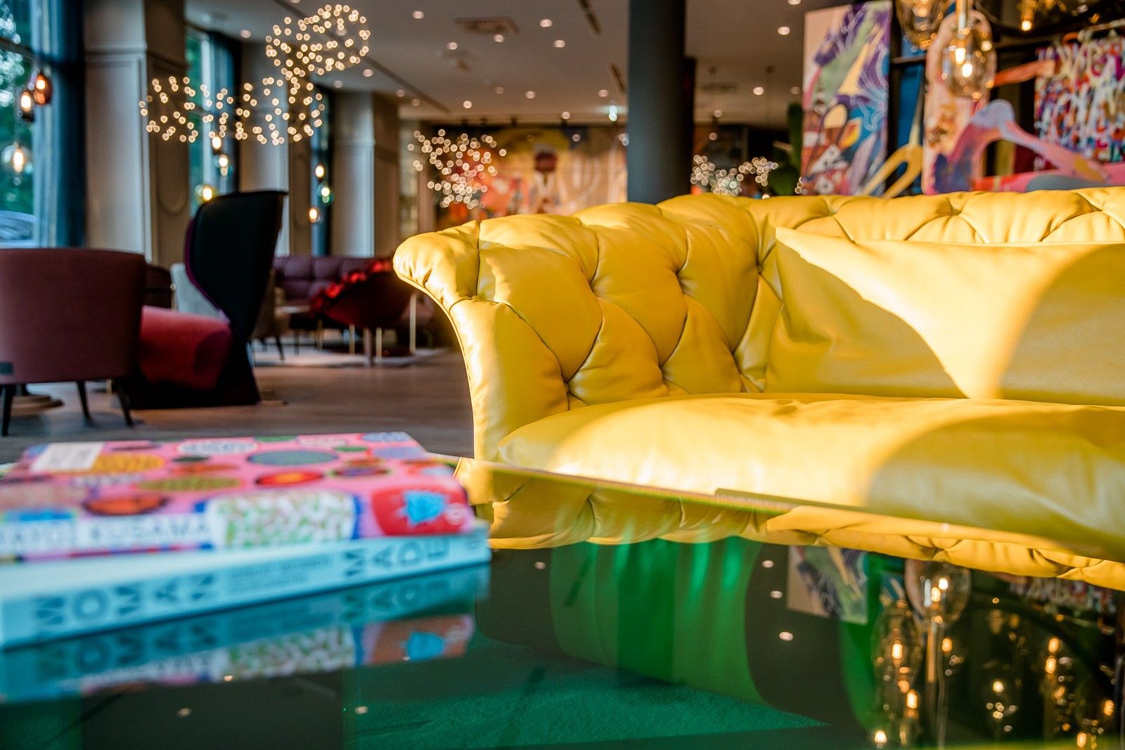 Gemütliche Lounge mit gelbem Chesterfield-Sofa, stilvollen Sesseln und moderner Beleuchtung. Farbenfrohe Kunst an den Wänden schafft eine einladende Atmosphäre für entspannte Stunden im Hotel.