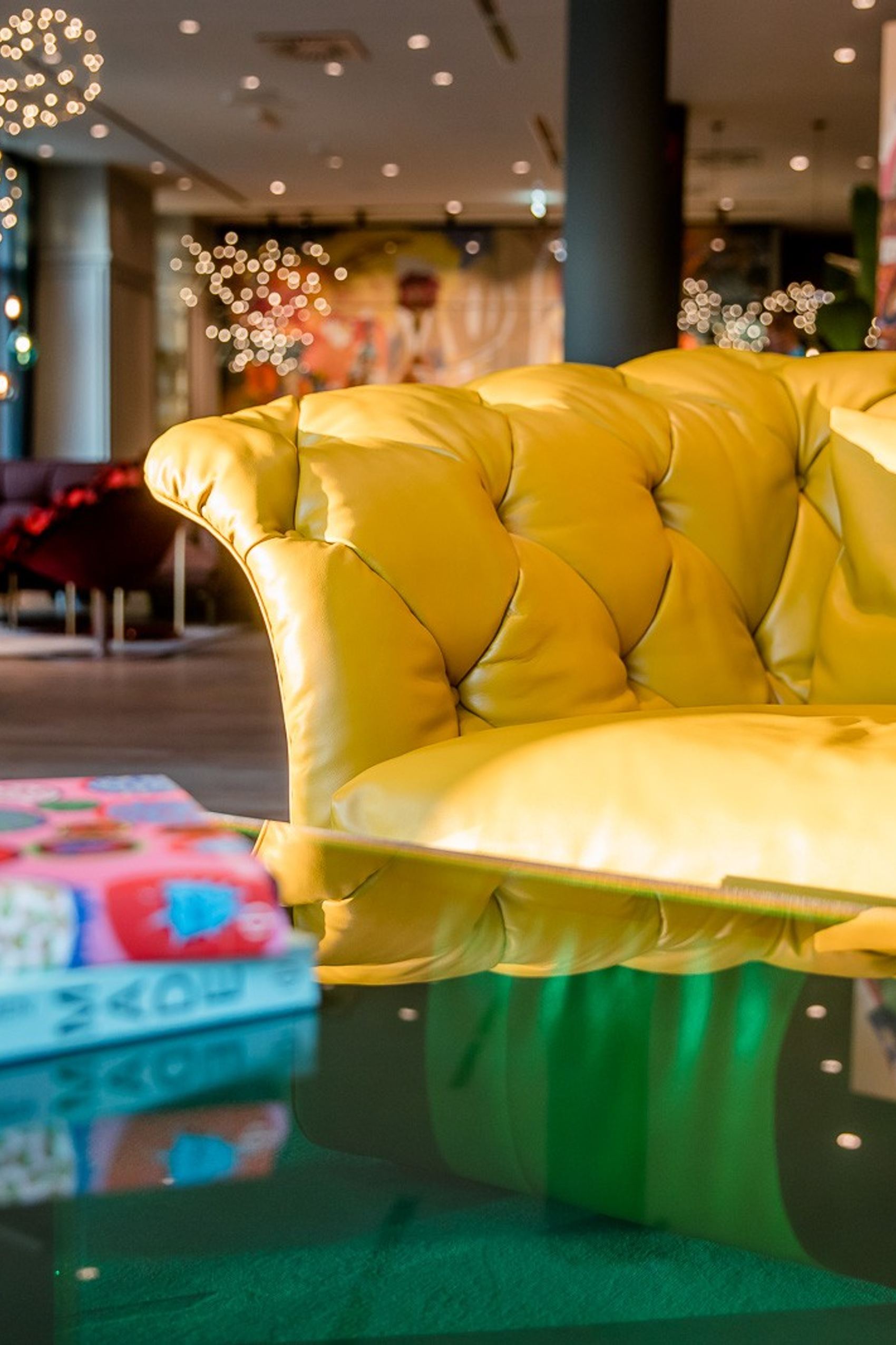 Gemütliche Lounge mit gelbem Chesterfield-Sofa, stilvollen Sesseln und moderner Beleuchtung. Farbenfrohe Kunst an den Wänden schafft eine einladende Atmosphäre für entspannte Stunden im Hotel.