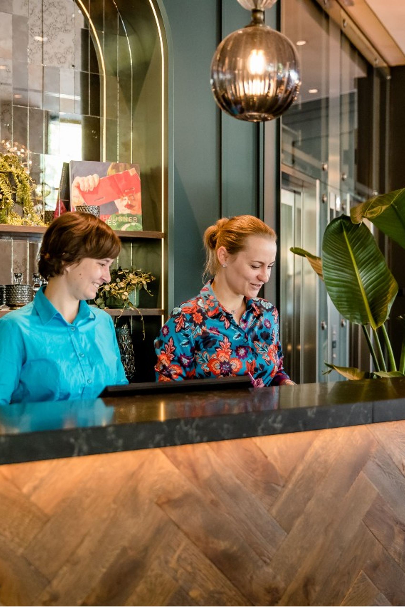 Zwei freundlich lächelnde Mitarbeiterinnen begrüßen Gäste an einer eleganten Hotelrezeption mit stilvollen Lampen und Pflanzen, die eine einladende und moderne Atmosphäre schaffen.