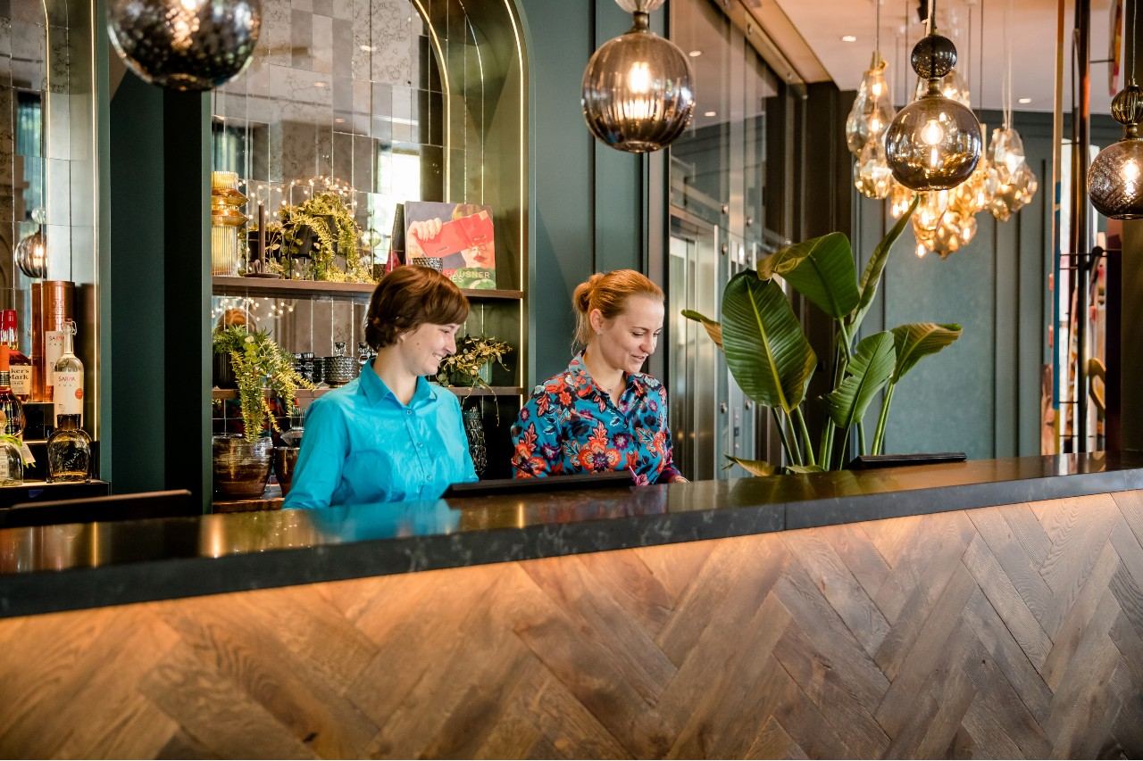 Zwei freundlich lächelnde Mitarbeiterinnen begrüßen Gäste an einer eleganten Hotelrezeption mit stilvollen Lampen und Pflanzen, die eine einladende und moderne Atmosphäre schaffen. Zwei freundlich lächelnde Mitarbeiterinnen begrüßen Gäste an einer eleganten Hotelrezeption mit stilvollen Lampen und Pflanzen, die eine einladende und moderne Atmosphäre schaffen.