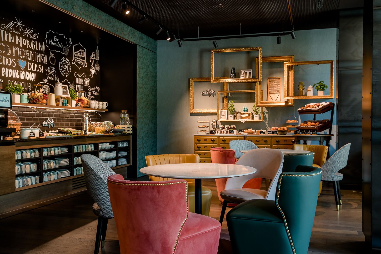 Gemütlicher Frühstücksbereich mit bunten Sesseln, runden Tischen und einem reichhaltigen Buffet mit Backwaren und Kaffeeangebot, eingerahmt von dekorativen Bilderrahmen und warmem Licht.