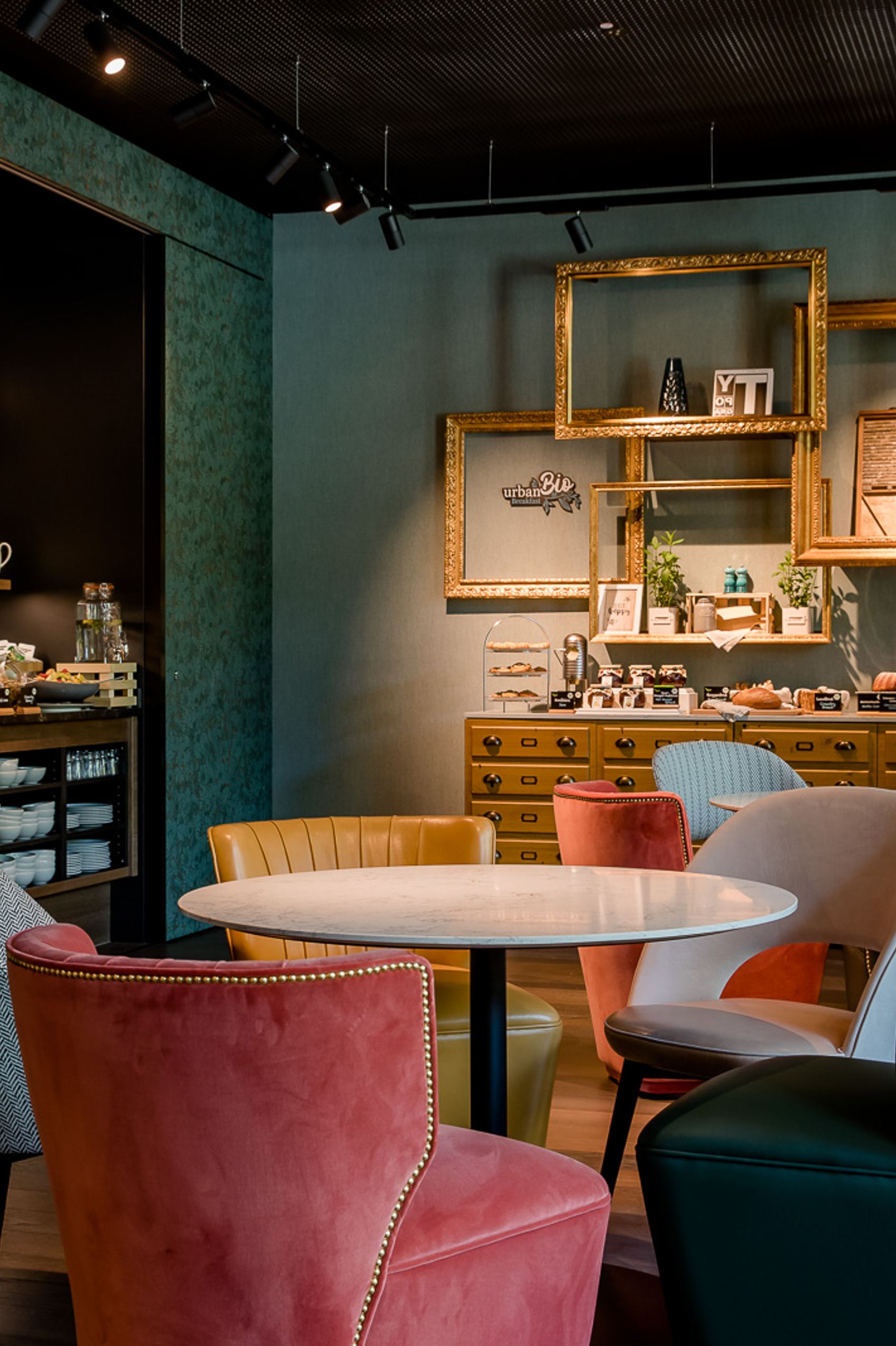 Gemütlicher Frühstücksbereich mit bunten Sesseln, runden Tischen und einem reichhaltigen Buffet mit Backwaren und Kaffeeangebot, eingerahmt von dekorativen Bilderrahmen und warmem Licht.