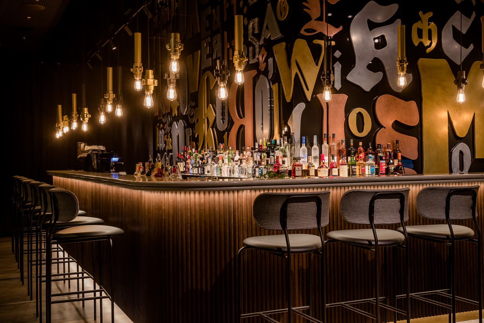 Moderne Hotelbar mit stilvoll beleuchtetem Tresen, hohen Barhockern und einer großen Auswahl an Spirituosen vor einer dekorativen Wand mit großformatigen Buchstaben. Warme, einladende Atmosphäre für entspannte Abende.