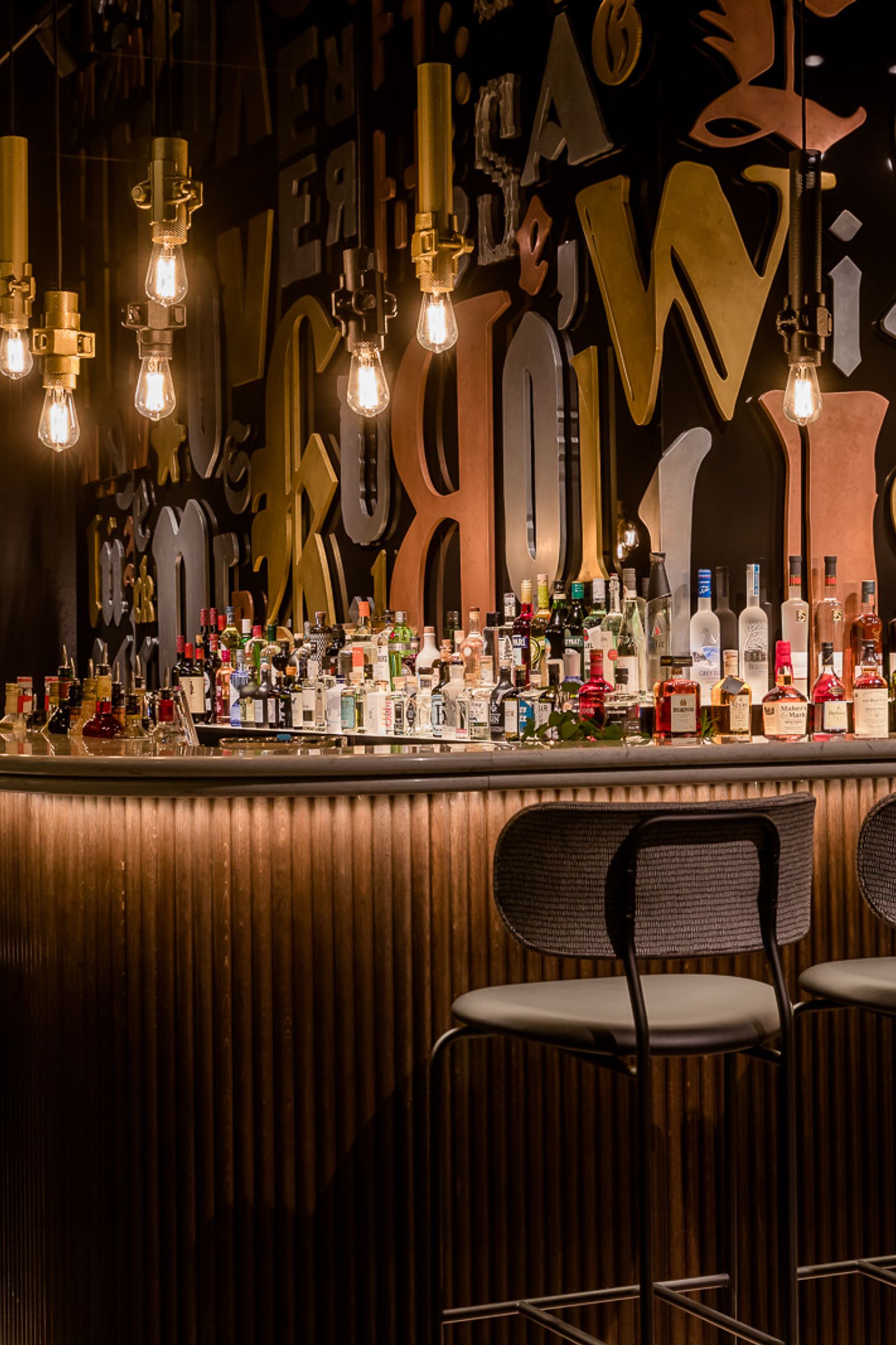 Motel One Hotelbar mit stilvoll beleuchtetem Tresen, hohen Barhockern und einer großen Auswahl an Spirituosen vor einer dekorativen Wand mit großformatigen Buchstaben.