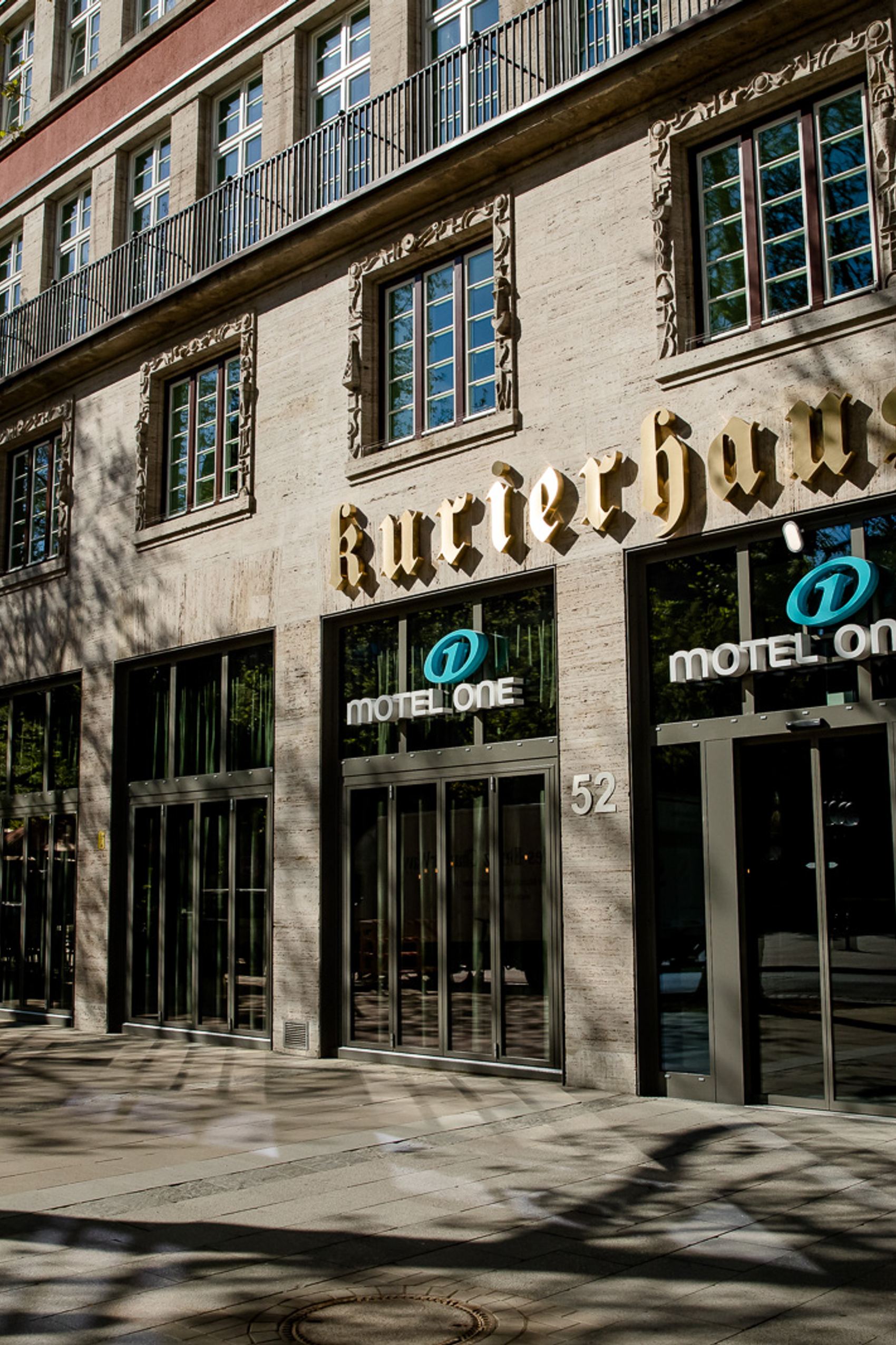 Fassade des Motel One Hotels in einem historischen Gebäude mit großen Fenstern und dem Schriftzug „Kurierhaus“ bei Tageslicht.