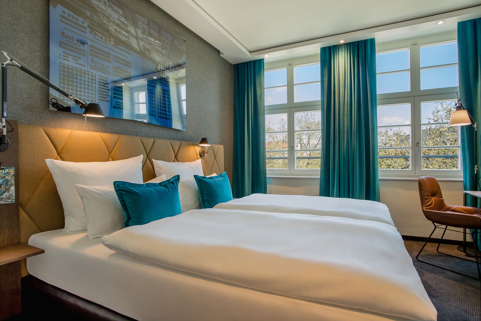 Helles Hotelzimmer mit großem Doppelbett, weißer Bettwäsche und türkisfarbenen Kissen. Große Fenster mit Ausblick und passenden türkisfarbenen Vorhängen sorgen für eine freundliche Atmosphäre. Modernes, gemütliches Design mit Schreibtisch und angenehmer Beleuchtung.