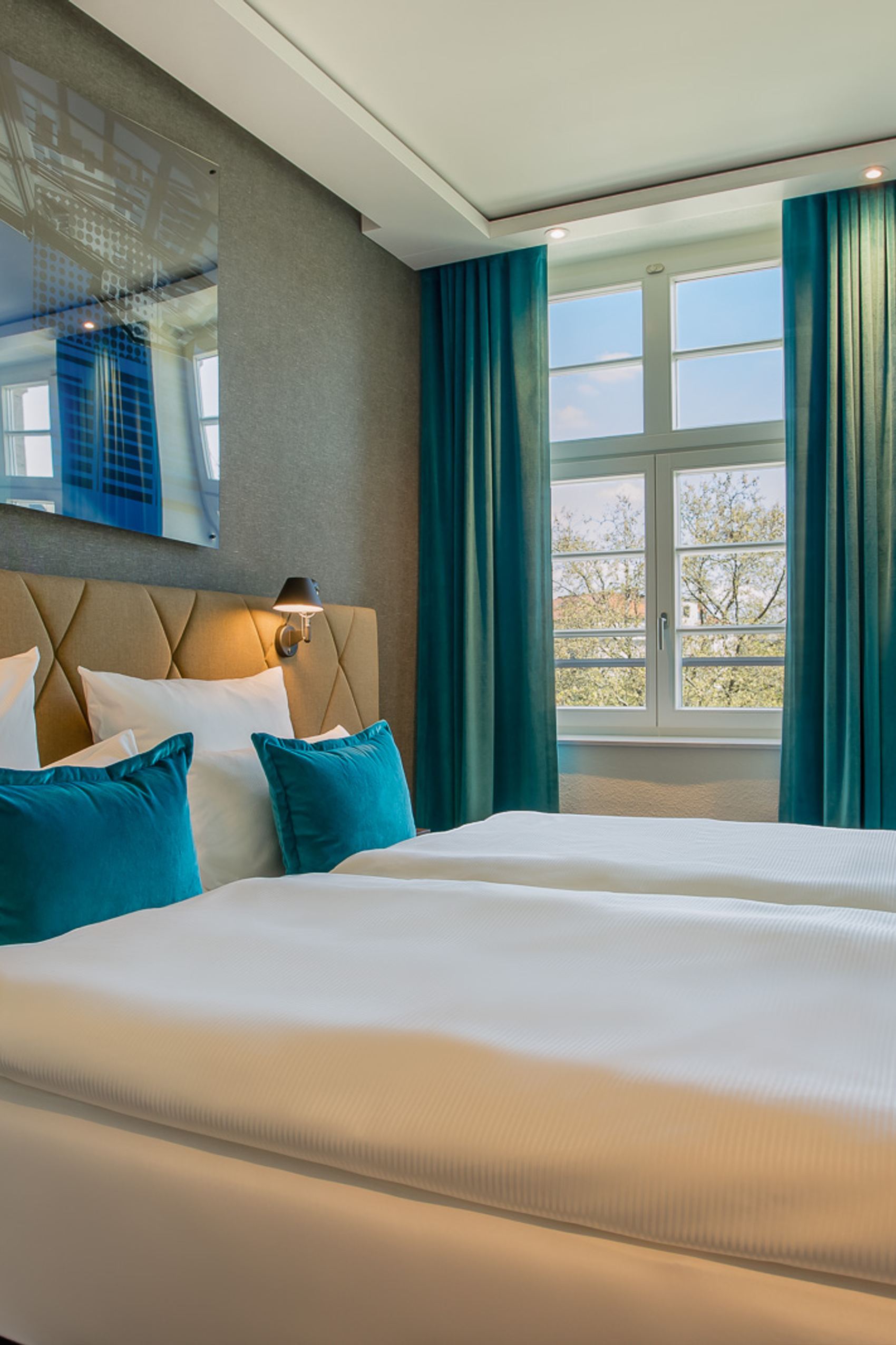 Helles Hotelzimmer mit großem Doppelbett, weißer Bettwäsche und türkisfarbenen Kissen. Große Fenster mit Ausblick und passenden türkisfarbenen Vorhängen sorgen für eine freundliche Atmosphäre. Modernes, gemütliches Design mit Schreibtisch und angenehmer Beleuchtung.