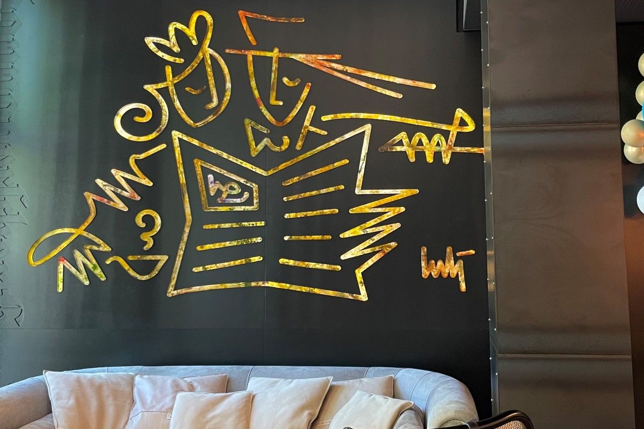 Goldene, abstrakte Wandkunst über einem hellen Sofa, die eine stilisierte Szenerie mit Figuren und einem geöffneten Buch darstellt.