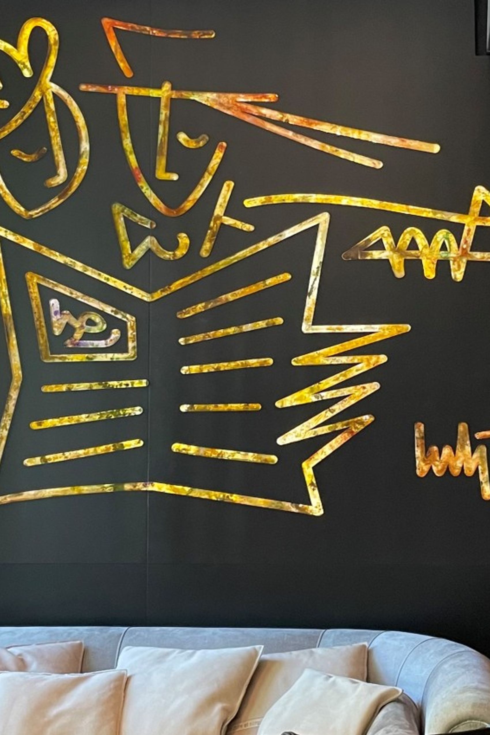Goldene, abstrakte Wandkunst über einem hellen Sofa, die eine stilisierte Szenerie mit Figuren und einem geöffneten Buch darstellt.