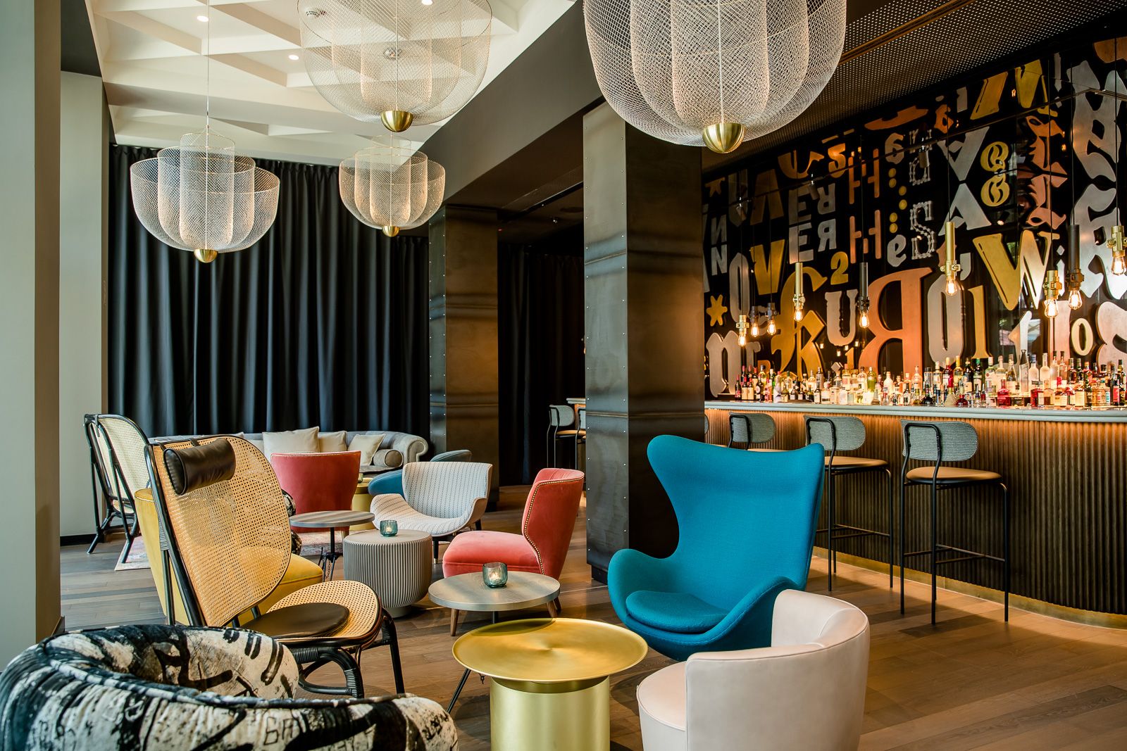 Moderne Hotelbar mit farbenfrohen, komfortablen Sesseln, stilvollen goldenen Beistelltischen und einer beleuchteten Bar mit dekorativen Buchstaben an der Wand. Großzügige Sitzbereiche laden zum Verweilen und Entspannen ein. 