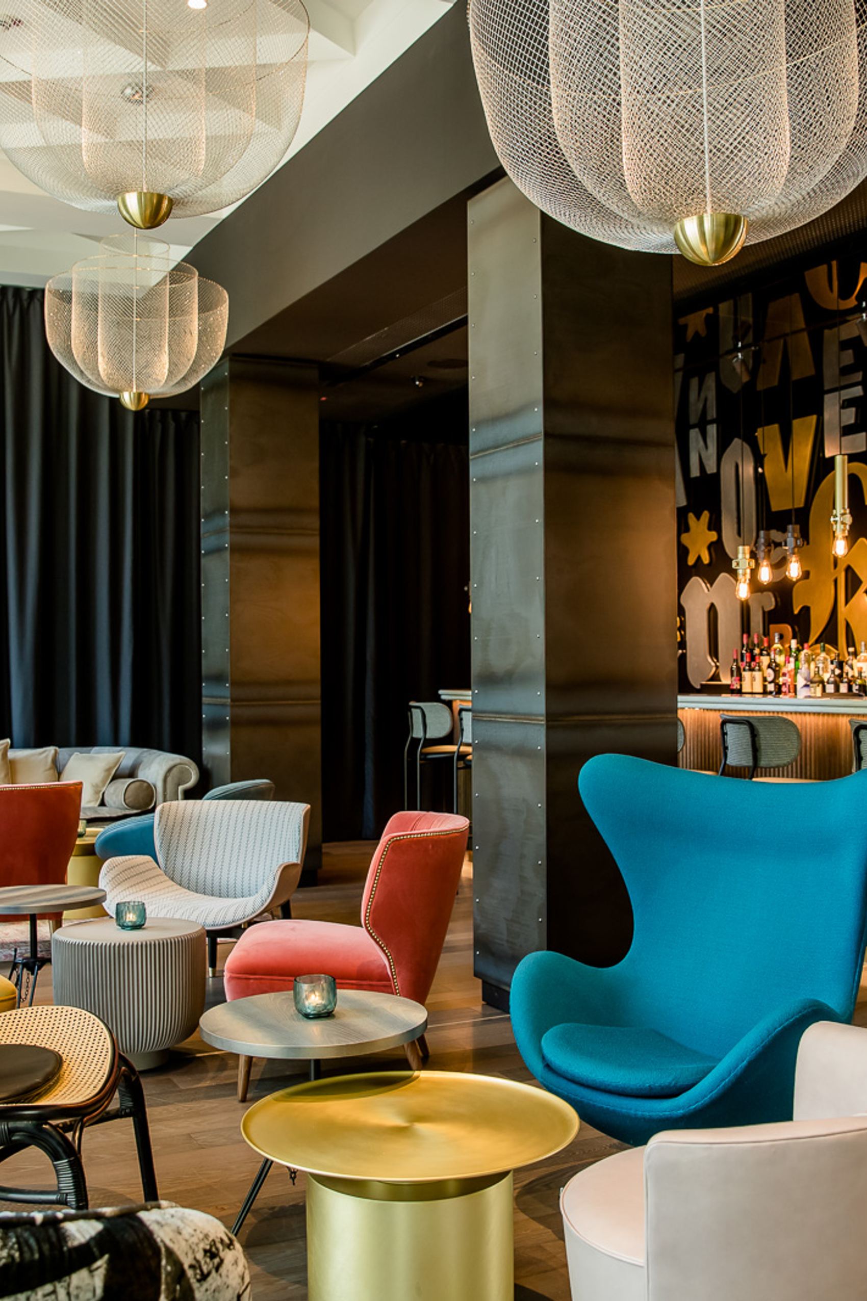 Motel One Hotelbar mit farbenfrohen, komfortablen Sesseln, stilvollen goldenen Beistelltischen und einer beleuchteten Bar mit dekorativen Buchstaben an der Wand. Großzügige Sitzbereiche laden zum Verweilen und Entspannen ein.