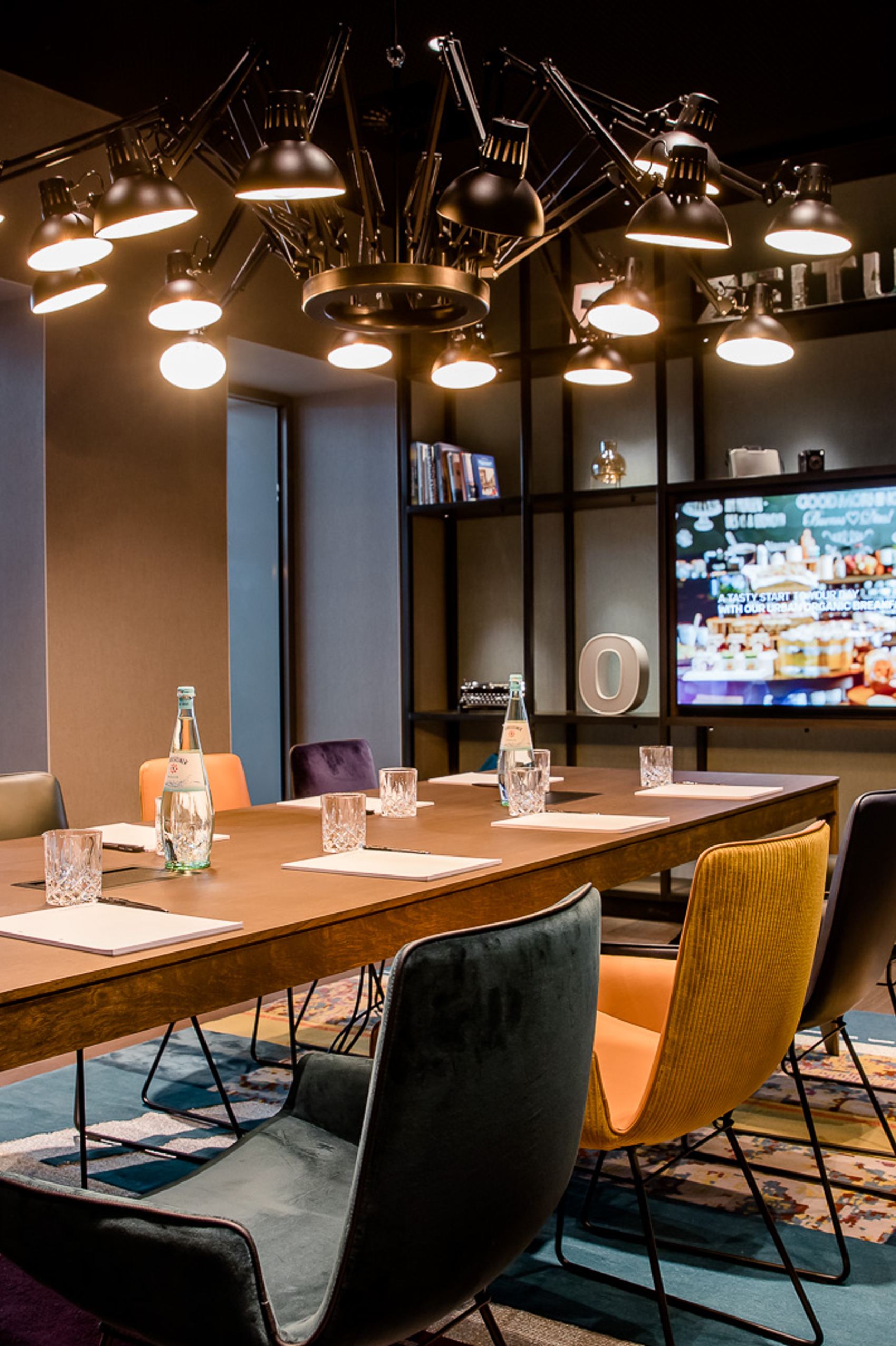 Motel One Konferenzraum mit großem Holztisch, verschiedenen bunten Stühlen, Wasserflaschen und Gläsern, stilvoller Beleuchtung und einem großen Bildschirm für Präsentationen