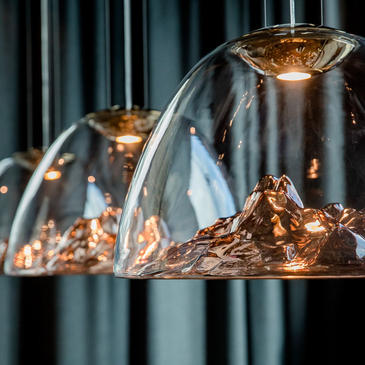 Design-Hängelampen mit glasklaren Kuppeln, in deren Inneren detailreiche Berglandschaften aus glänzendem Metall geformt sind, strahlen warmes Licht aus