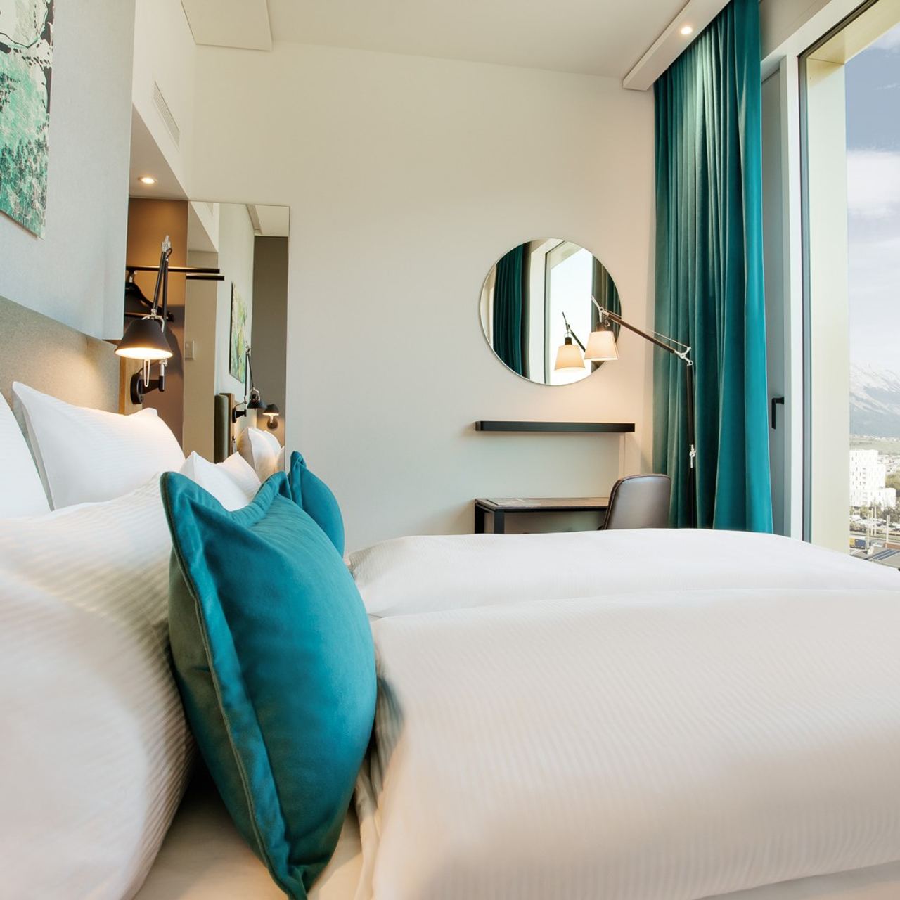 Hotelzimmer mit großem, bequemem Doppelbett, weißen Bettwäsche und türkisfarbenen Kissen, hellem Arbeitsbereich mit runden Spiegel, großer Fensterfront.