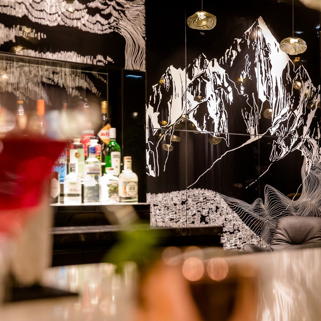 Blick auf eine stilvoll beleuchtete Bar mit einer Auswahl an Spirituosen vor einer schwarzen Wand, die mit künstlerischer Berglandschaft in Weiß verziert ist. Die Atmosphäre wirkt modern und gemütlich.