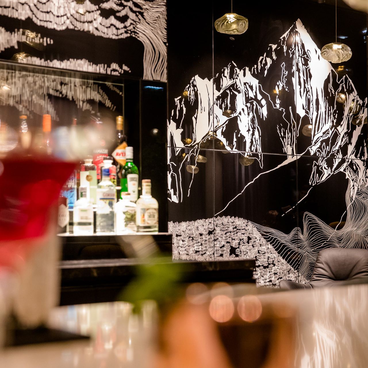 Blick auf eine stilvoll beleuchtete Bar mit einer Auswahl an Spirituosen vor einer schwarzen Wand, die mit künstlerischer Berglandschaft in Weiß verziert ist. Die Atmosphäre wirkt modern und gemütlich.