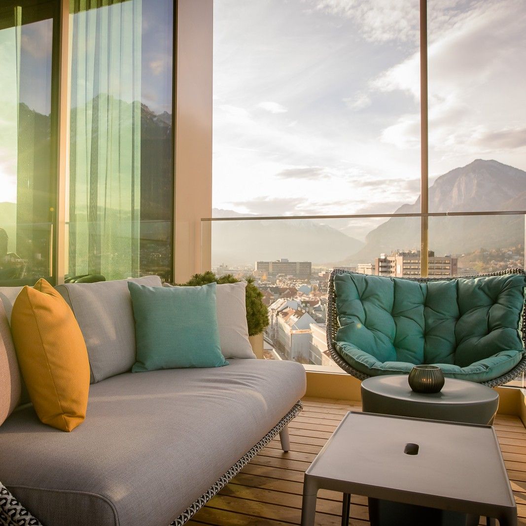 Moderne Lounge mit gemütlichem Sofa und Sessel auf Balkon, große Fenster bieten Panorama auf Stadt und Berge bei Sonnenuntergang.