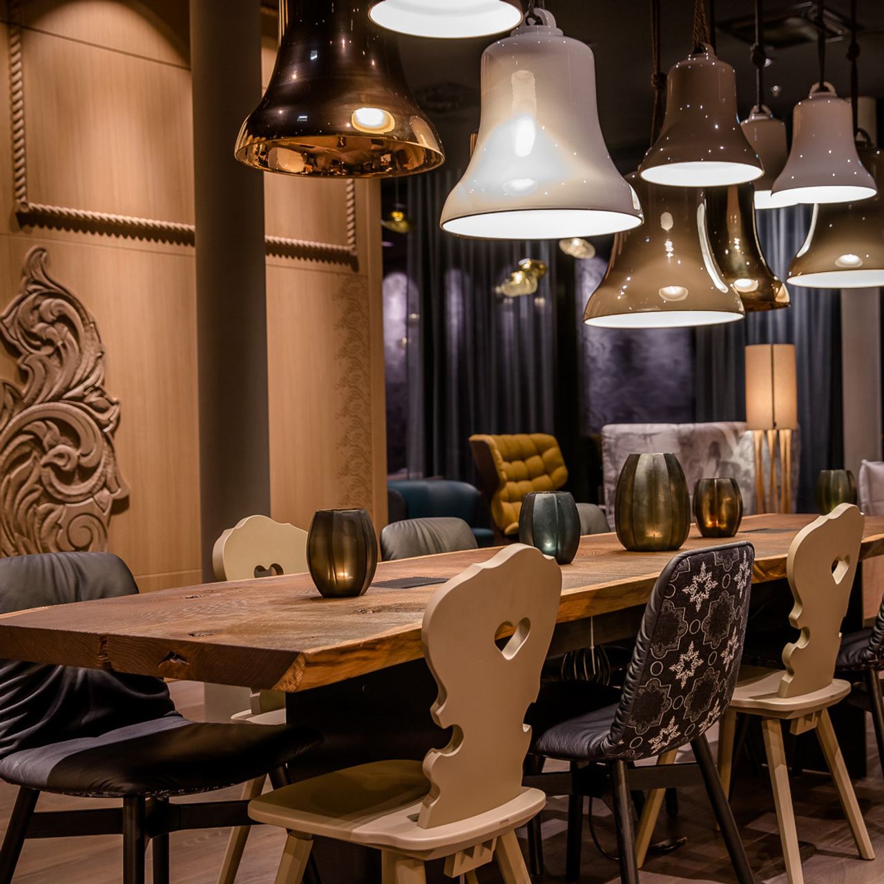 Gemütlicher Essbereich mit rustikalem Holztisch, verschiedenen Stühlen und mehreren modernen, unterschiedlich geformten Pendelleuchten, die warmes Licht spenden. Im Hintergrund gemütliche Sitzmöbel und dekorative Wandverkleidung schaffen eine einladende Atmosphäre.