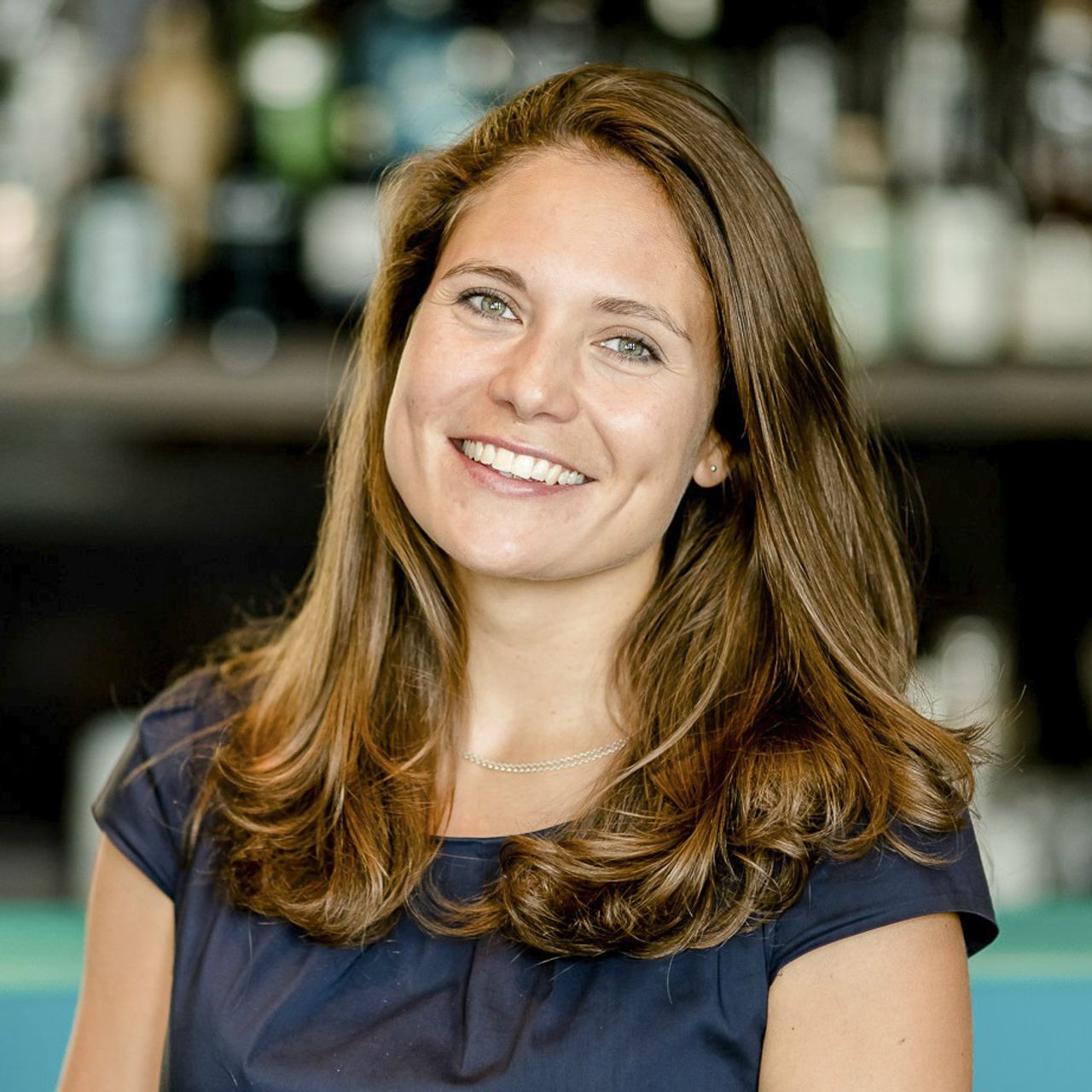 Lächelnde junge Frau mit schulterlangem braunem Haar, die freundlich in die Kamera blickt, vor unscharfem Hintergrund.