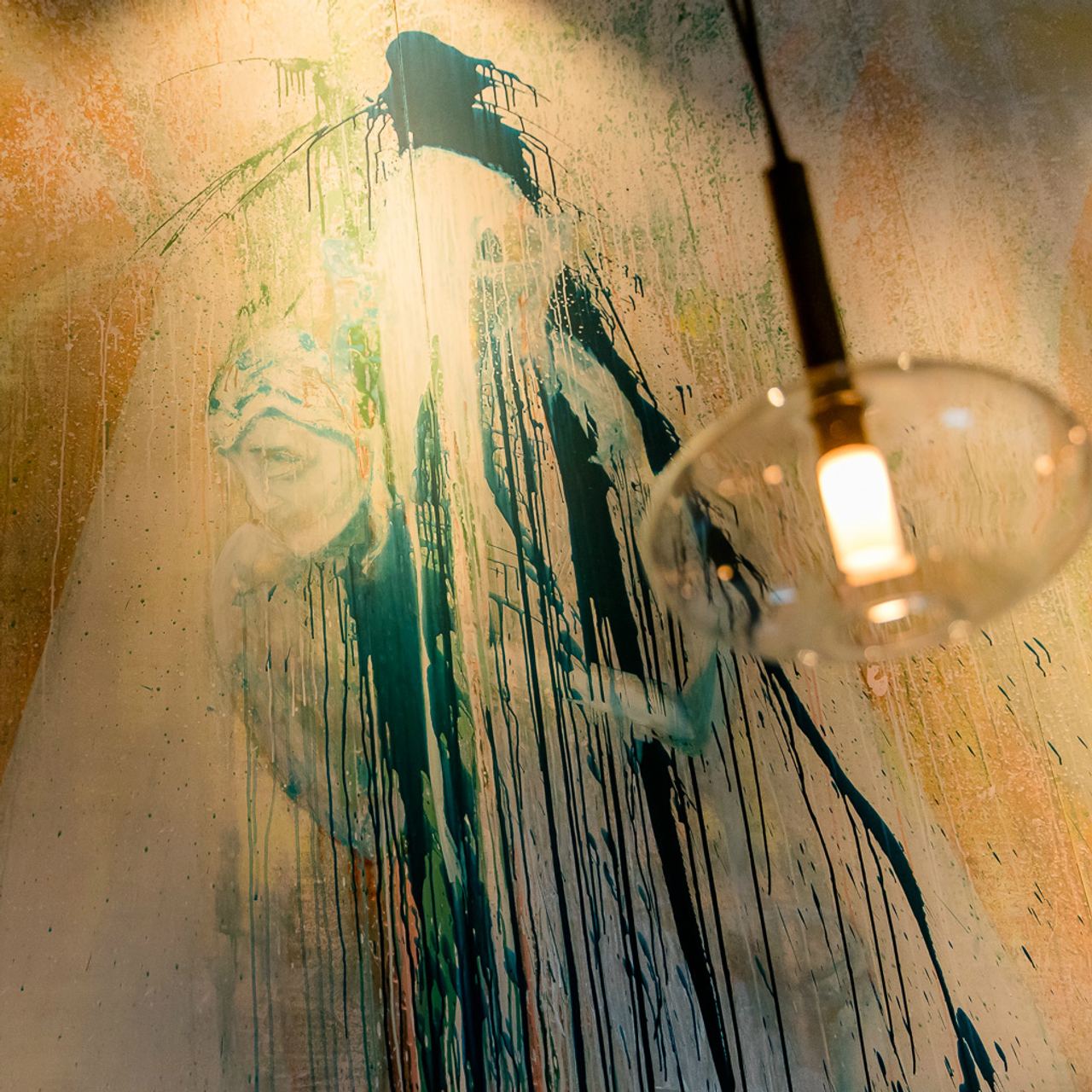 Wandkunst mit fließenden Farbspuren in Blau- und Grüntönen, die ein abstraktes Gesicht und eine Figur andeuten; im Vordergrund eine moderne, schwebende Hängelampe mit warmem Licht.