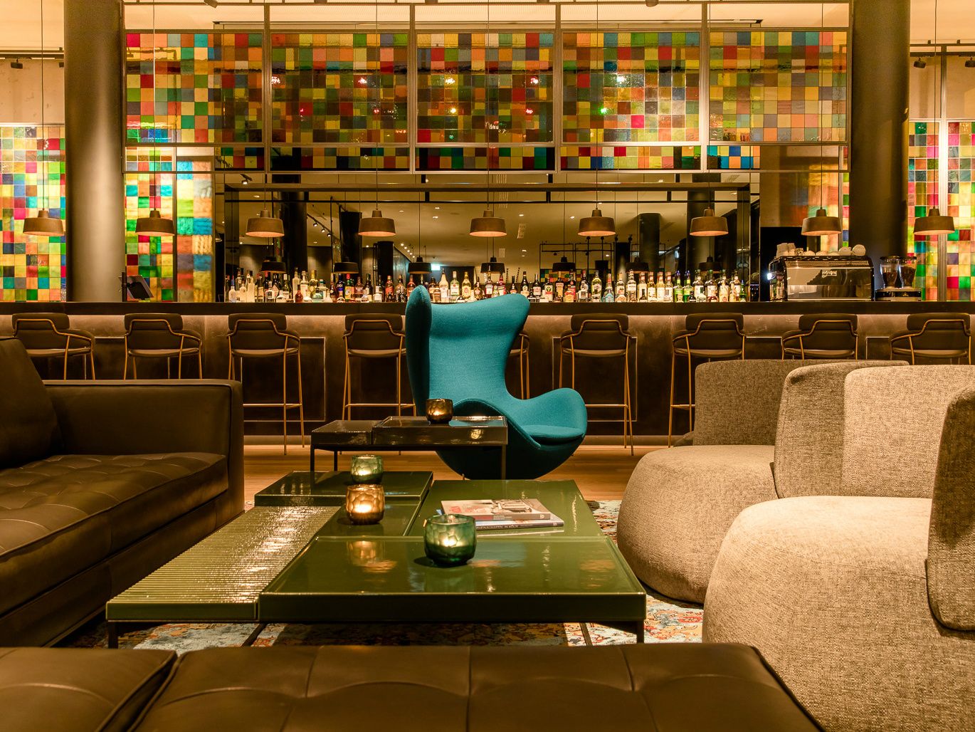 Moderne Hotelbar mit stilvoller Sitzgruppe aus Sesseln und Sofas, farbenfroher Mosaikwand und gedämpfter Beleuchtung.