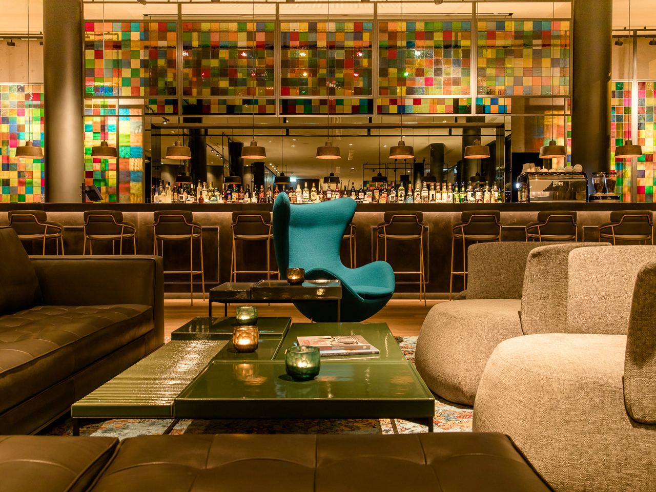 Motel One Hotelbar mit stilvoller Sitzgruppe aus Sesseln und Sofas, farbenfroher Mosaikwand und gedämpfter Beleuchtung.