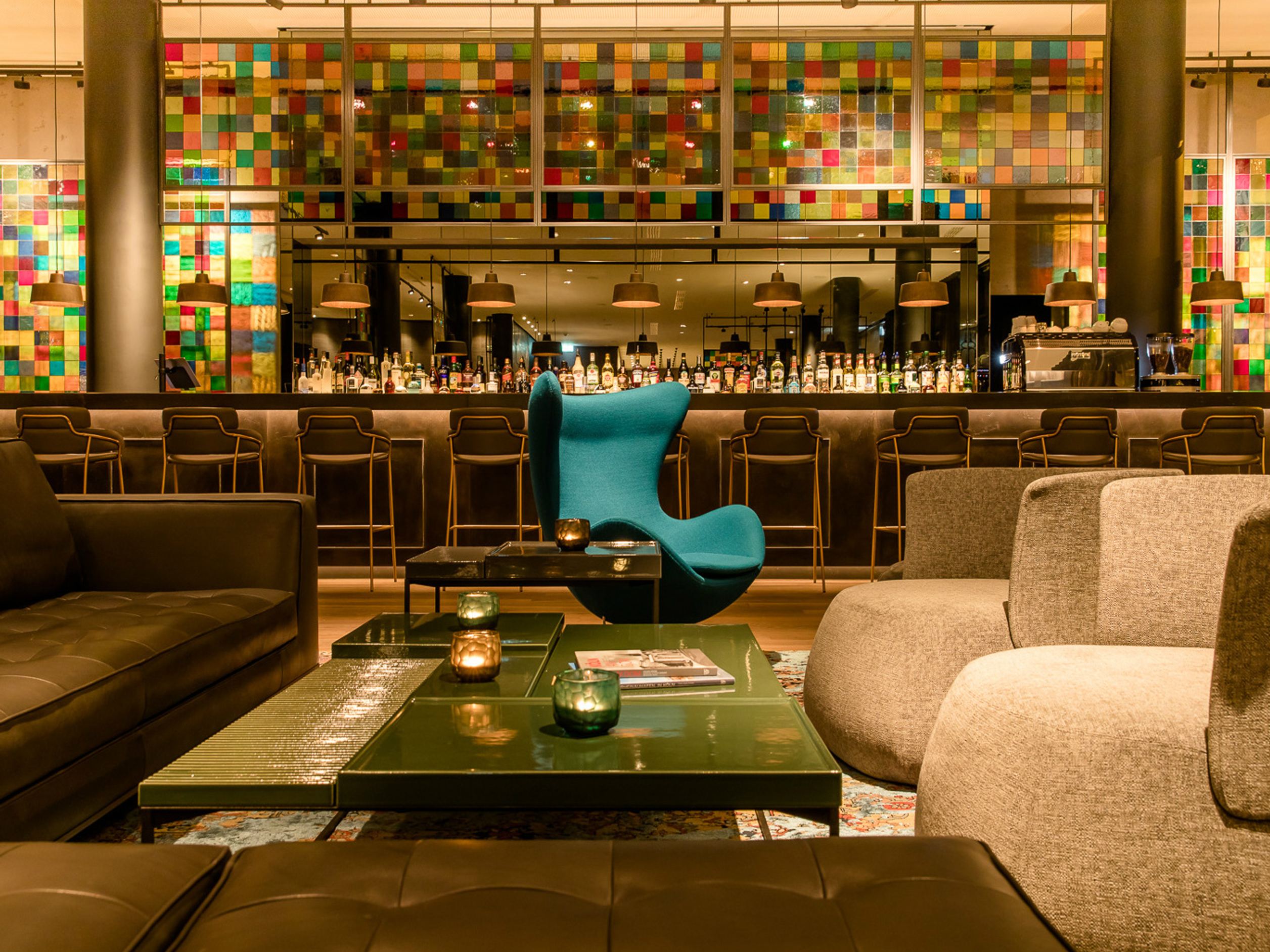 Motel One Hotelbar mit stilvoller Sitzgruppe aus Sesseln und Sofas, farbenfroher Mosaikwand und gedämpfter Beleuchtung.