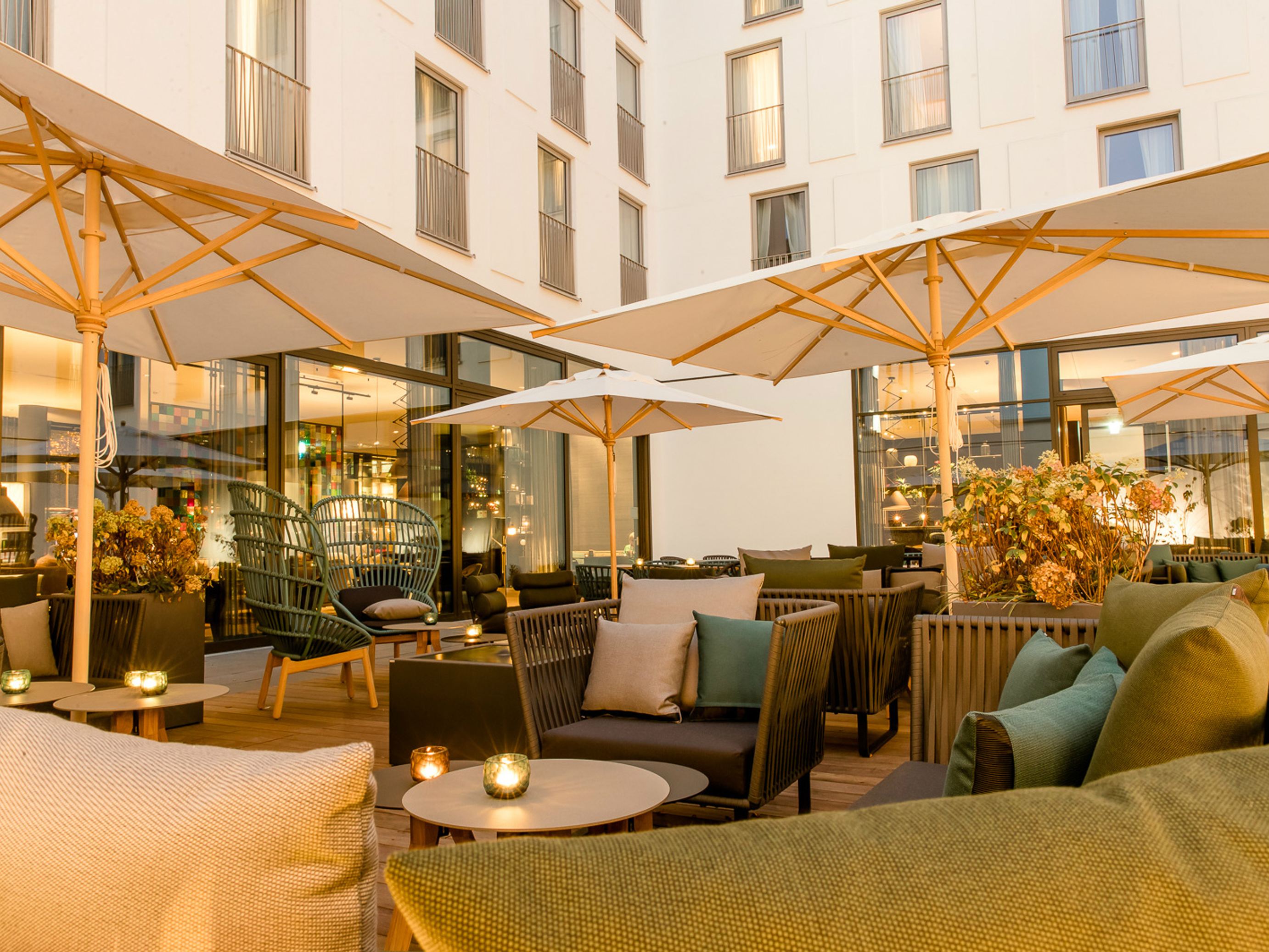 Motel One Außenbereich einer Hotelterrasse mit gemütlichen Sitzgruppen, weichen Kissen und großen Sonnenschirmen, stimmungsvolle Beleuchtung.