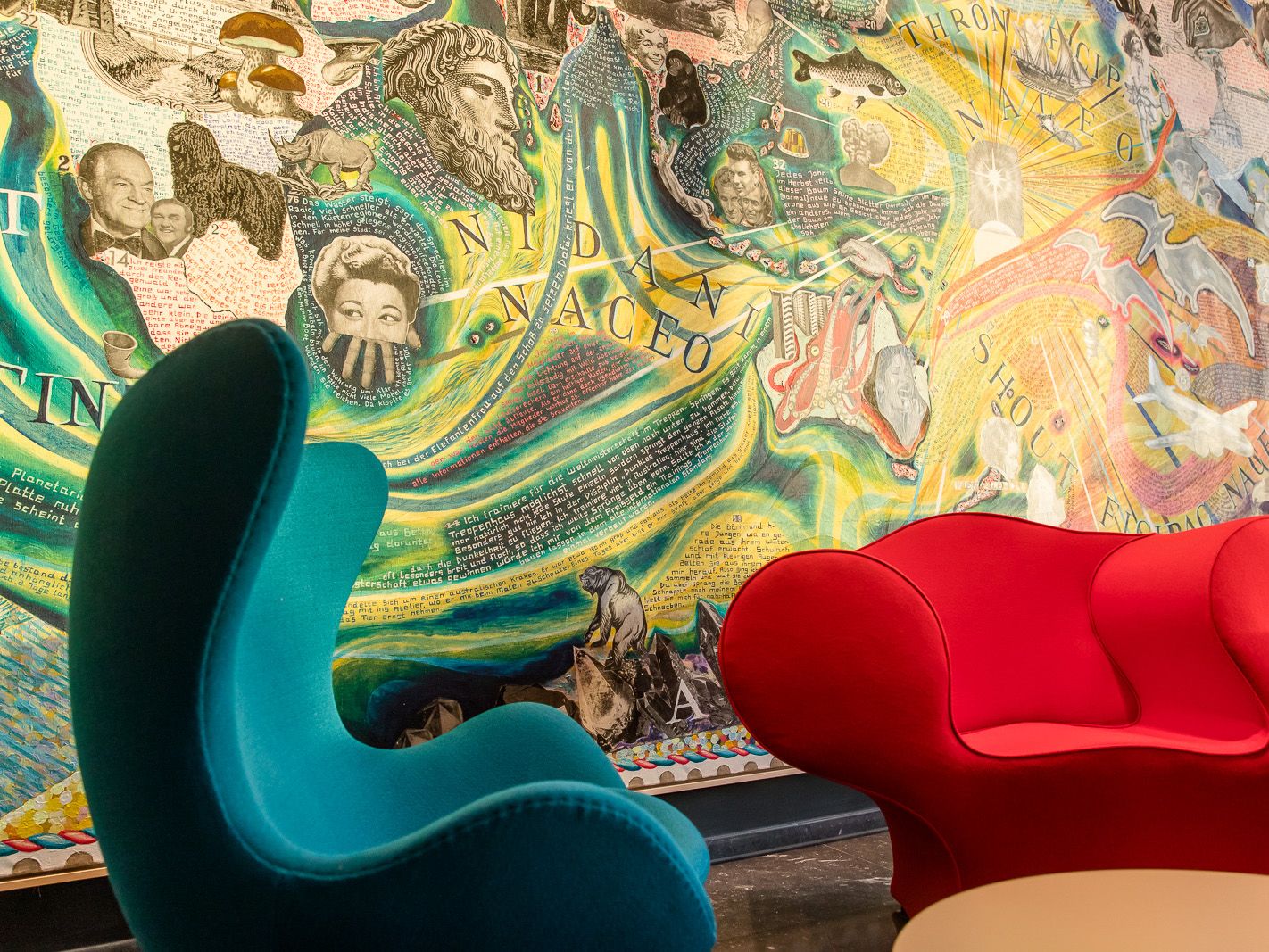 Zwei farbige Design-Sessel, einer in Blau und einer in Rot, vor einer lebendigen, detailreichen Wandmalerei mit Gesichtern, Schriftzügen und abstrakten Mustern. Die Szene strahlt kreative Atmosphäre und modernen Komfort aus.