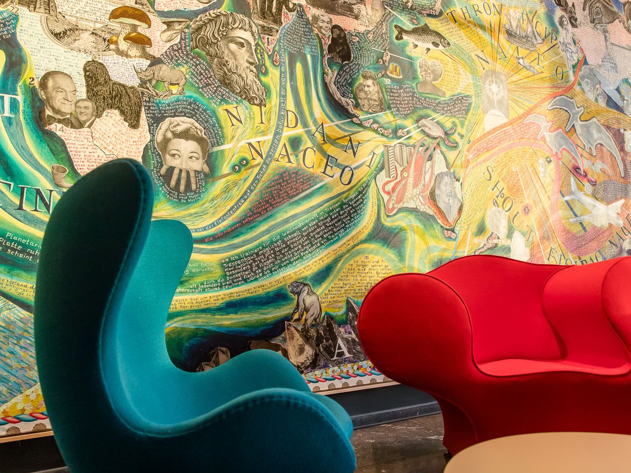 Zwei farbige Design-Sessel, einer in Blau und einer in Rot, vor einer lebendigen, detailreichen Wandmalerei mit Gesichtern, Schriftzügen und abstrakten Mustern. Die Szene strahlt kreative Atmosphäre und modernen Komfort aus.