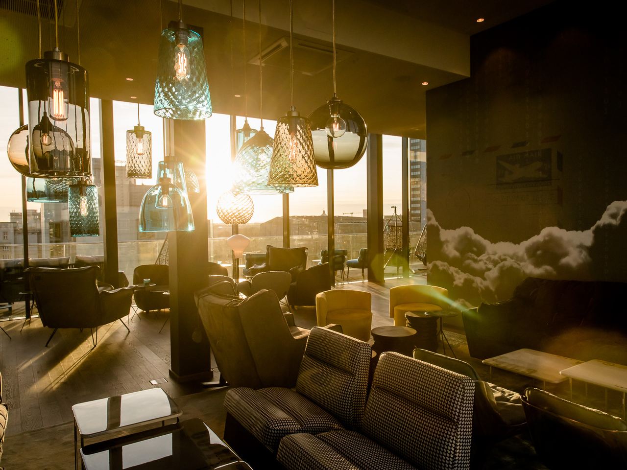 Gemütliche Motel One Hotel-Lounge mit bequemen Sesseln und Sofas, warmes Licht durch bunte Hängelampen, großer Fensterfront mit Blick auf die Stadt bei Sonnenuntergang