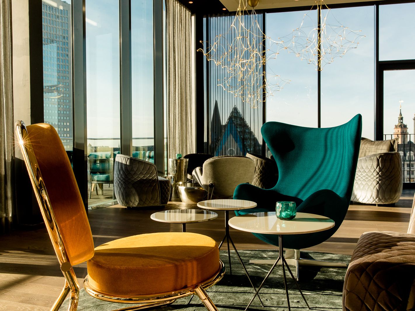 Lichtdurchfluteter Loungeraum mit modernen Sesseln und Tischen, großer Fensterfront mit Stadtblick, eleganter, entspannter Atmosphäre und stilvoller Beleuchtung.