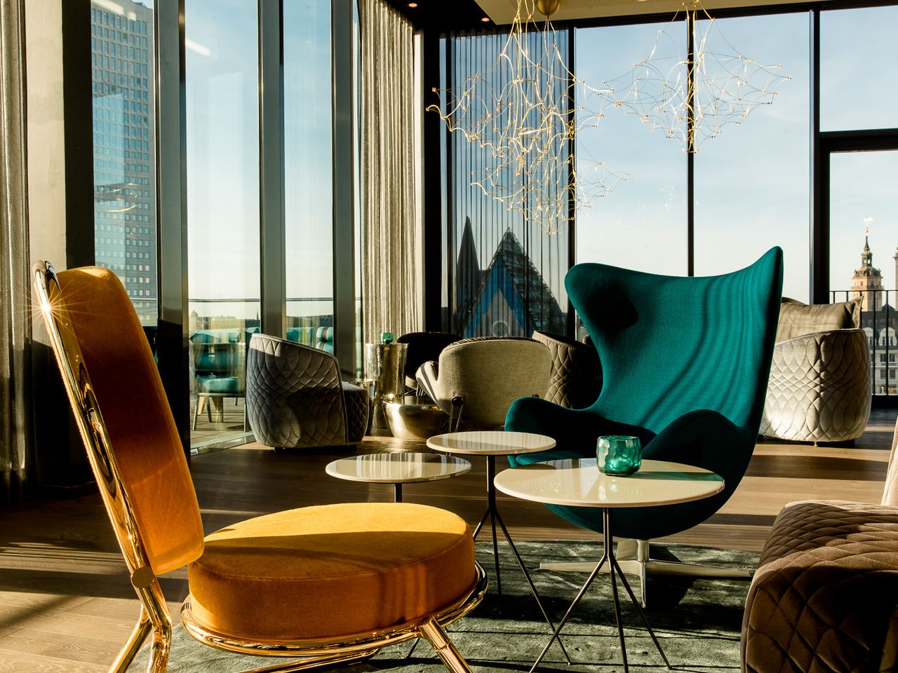 Lichtdurchfluteter Loungeraum mit modernen Sesseln und Tischen, großer Fensterfront mit Stadtblick, eleganter, entspannter Atmosphäre und stilvoller Beleuchtung.