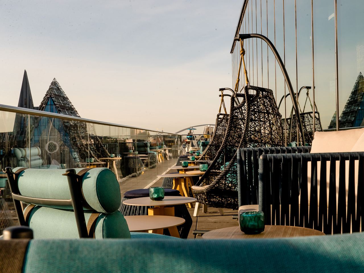 Dachterrasse mit stilvollen, gepolsterten Sitzmöbeln und Hängekörben, Tischen mit grünen Kerzenhaltern, umgeben von Glasgeländern, mit Blick auf die Stadt und den Himmel bei Sonnenuntergang.