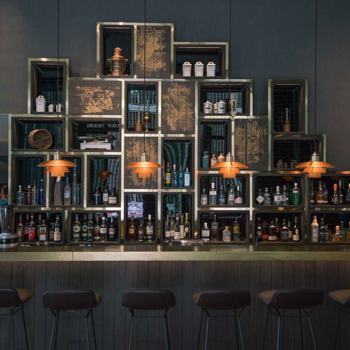 Bar mit Regal aus offenen Holzkästen, gefüllt mit verschiedenen Spirituosenflaschen und dekorativen Objekten, darunter Lampen mit warmem orangefarbenem Licht, gemütliche Barhocker davor.