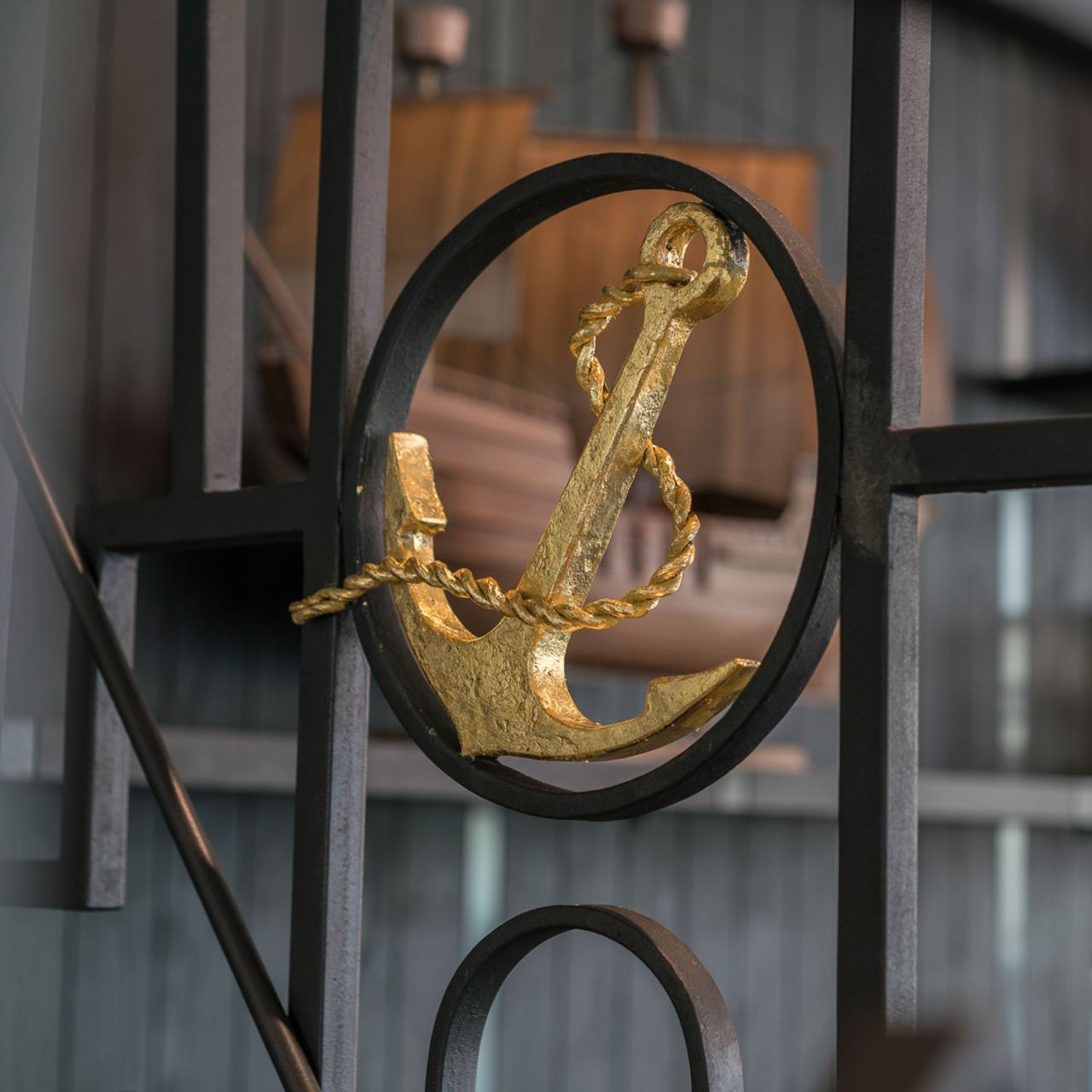 Goldener Anker mit Seil als dekoratives Symbol, eingebettet in einen schwarzen, runden Metallrahmen, vor unscharfem Hintergrund mit Holz- und Metallstrukturen.