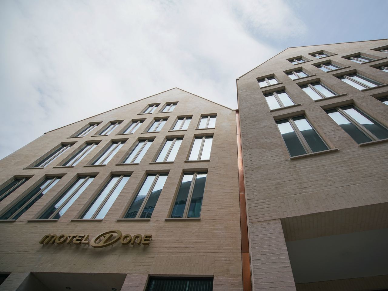 Helle Motel One Hotelgebäude mit großen Fensterfronten unter bewölktem Himmel, das Hotel-Logo ist an der Fassade sichtbar.