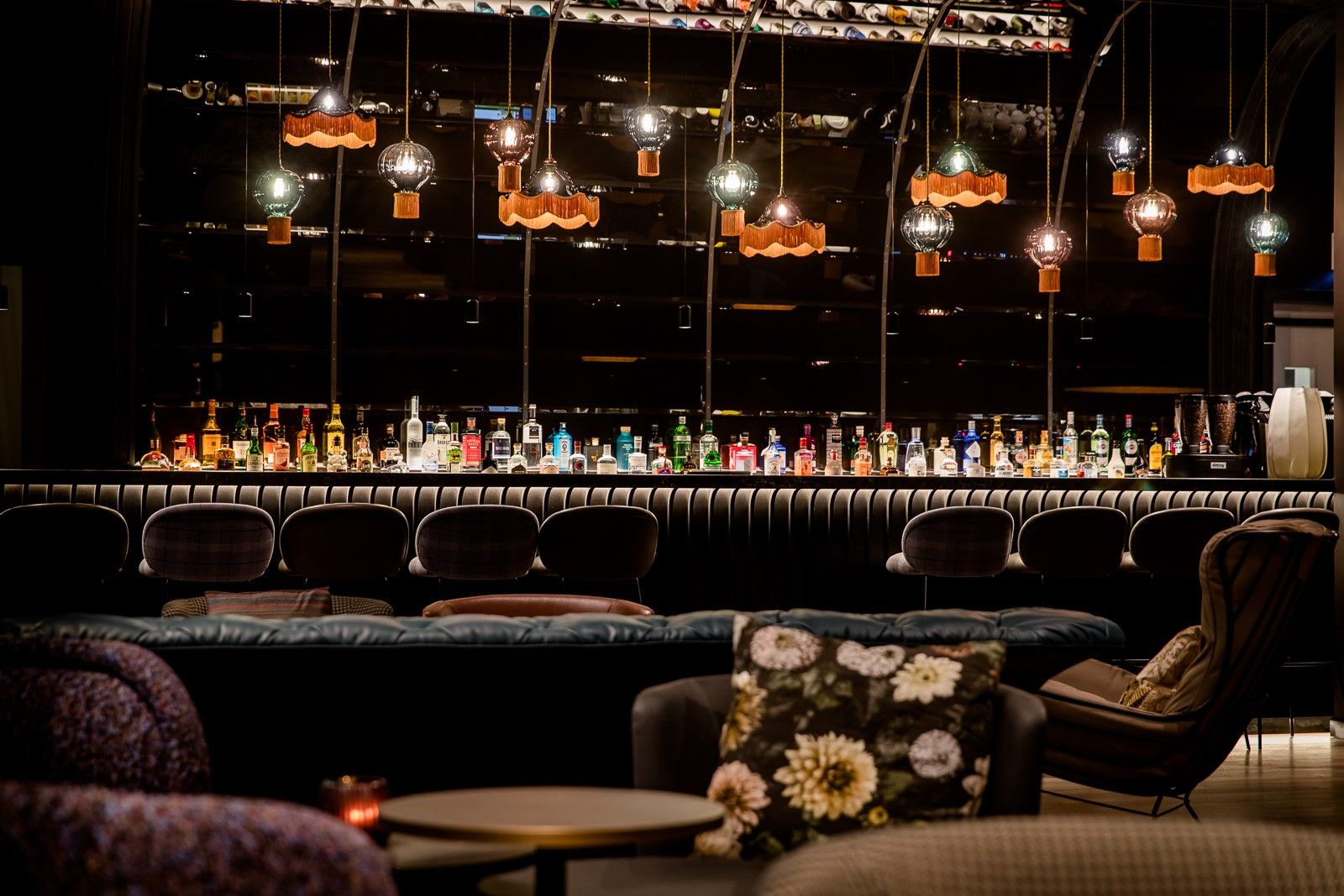 Moderne Bar mit einer langen Theke, darauf eine große Auswahl an bunten Spirituosenflaschen. Angenehme, warme Beleuchtung durch verschiedene hängende Lampen schafft eine einladende Atmosphäre zum Entspannen und Genießen. Gemütliche Sitzgelegenheiten mit Kissen im Vordergrund.