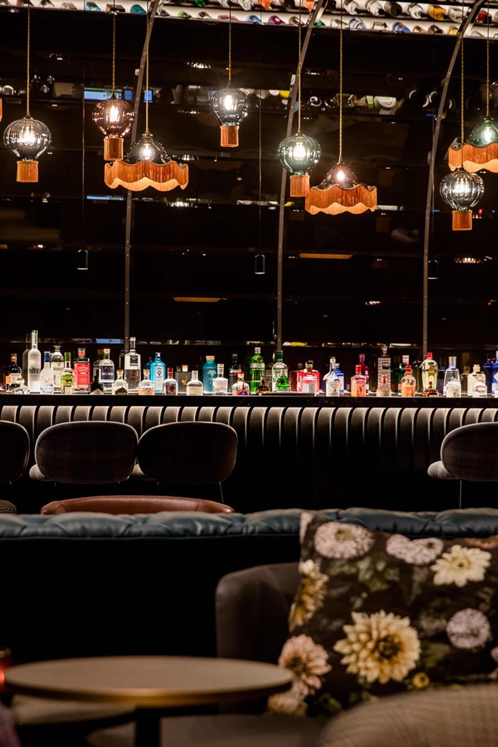 Motel One Bar mit einer langen Theke, darauf eine große Auswahl an bunten Spirituosenflaschen. Angenehme, warme Beleuchtung durch verschiedene hängende Lampen. Gemütliche Sitzgelegenheiten mit Kissen im Vordergrund.