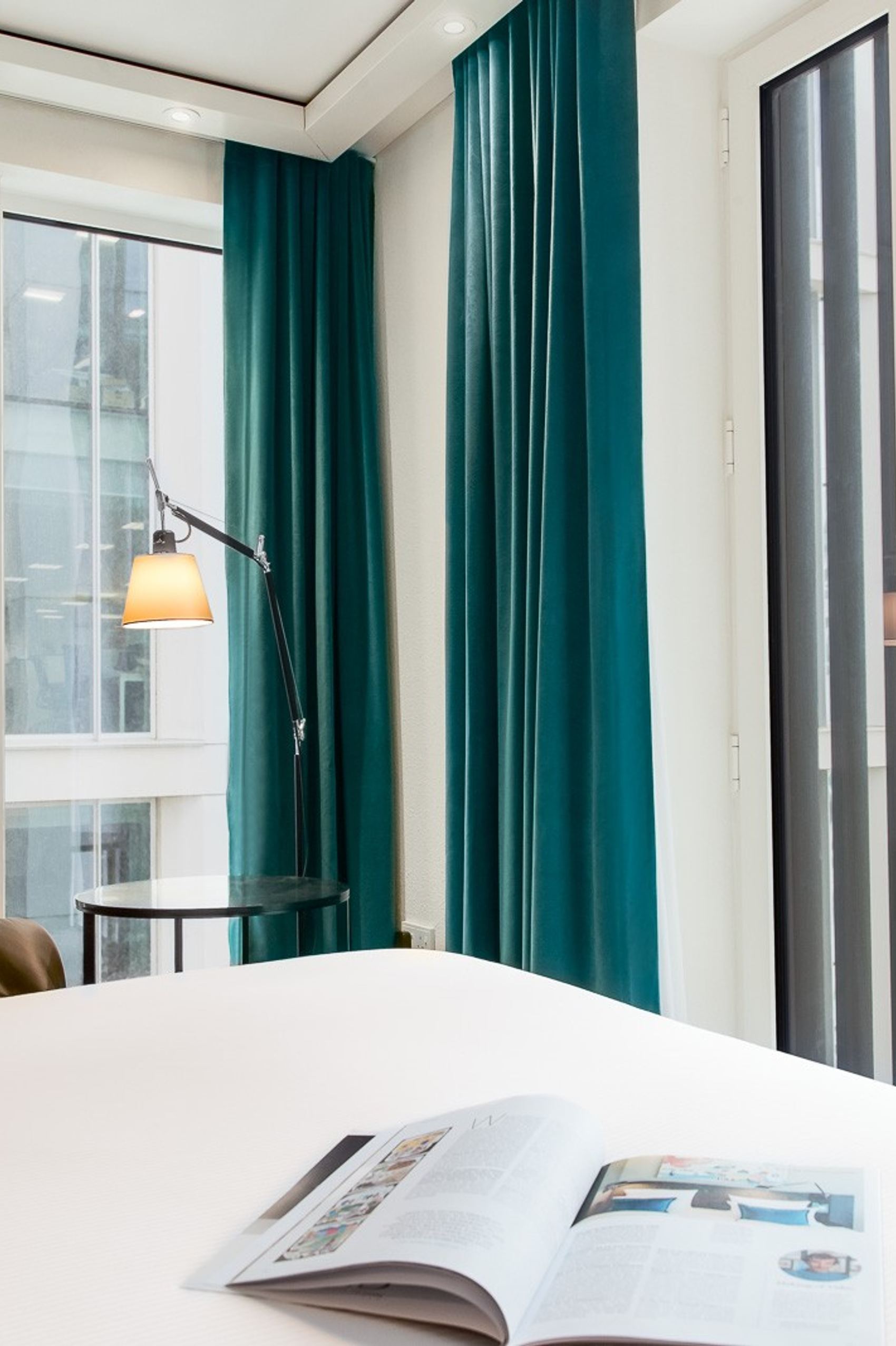 Helles Hotelzimmer mit großen Fenstern und blaugrünen Vorhängen, bequemer Ledersessel, Stehlampe und offenes Magazin auf dem Bett. Blick auf moderne und historische Gebäude in der Stadt.