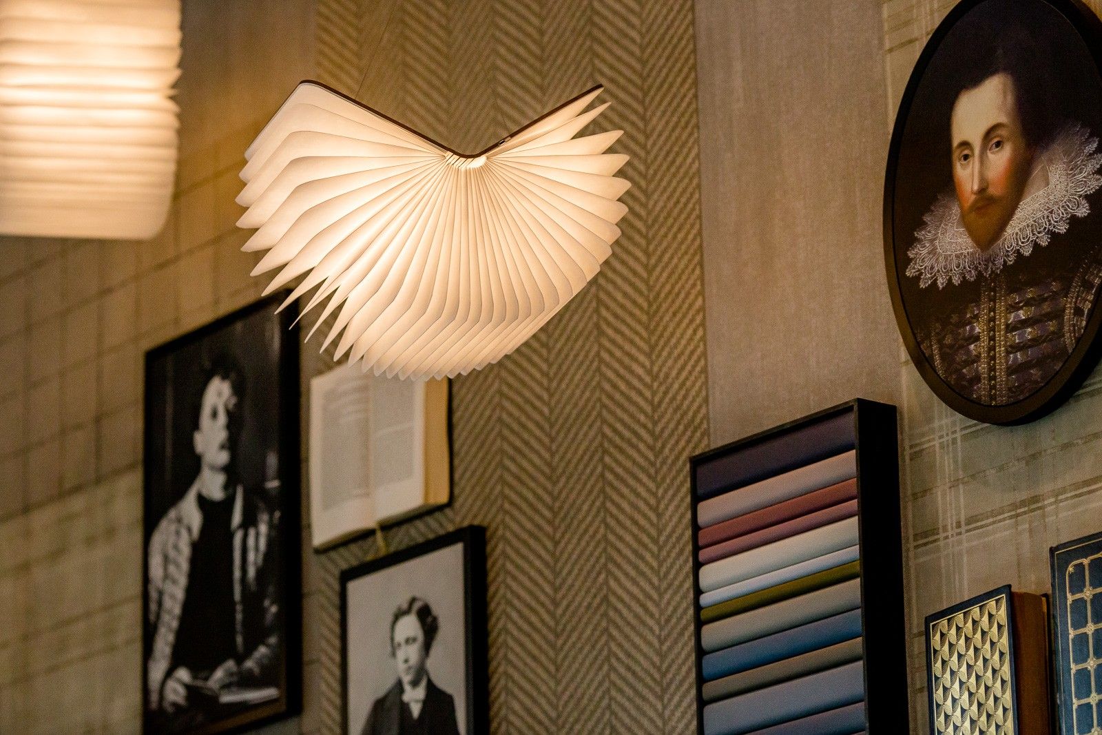 Dekorative Lampe in Bücherform an Wand mit klassischen Porträtbildern und Farbmustern.