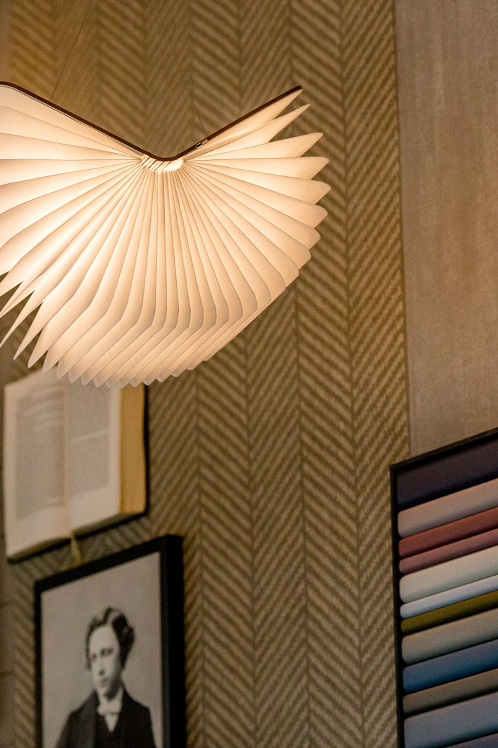 Dekorative Lampe in Bücherform an Wand mit klassischen Porträtbildern und Farbmustern.