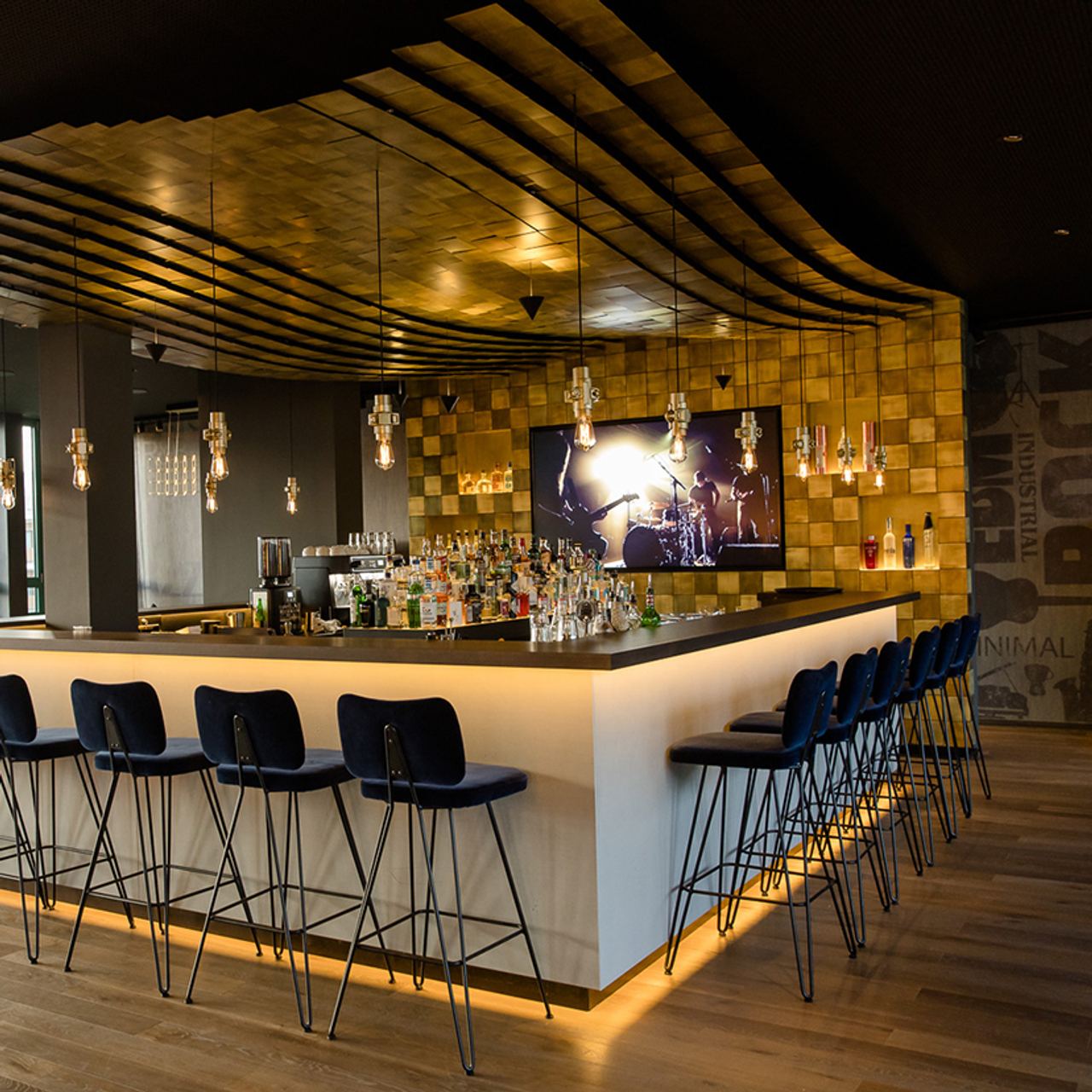 Motel One Hotelbar mit elegantem, beleuchtetem Tresen, stilvollen Hockern und warmem, wellenförmigem Deckendesign