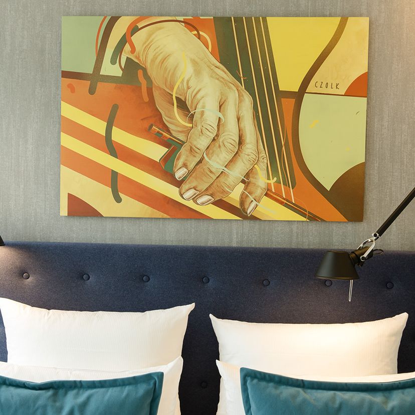 Motel One Gemälde über dem Bett zeigt eine Hand, die die Saiten eines Streichinstruments spielt, in warmen Orange- und Gelbtönen.