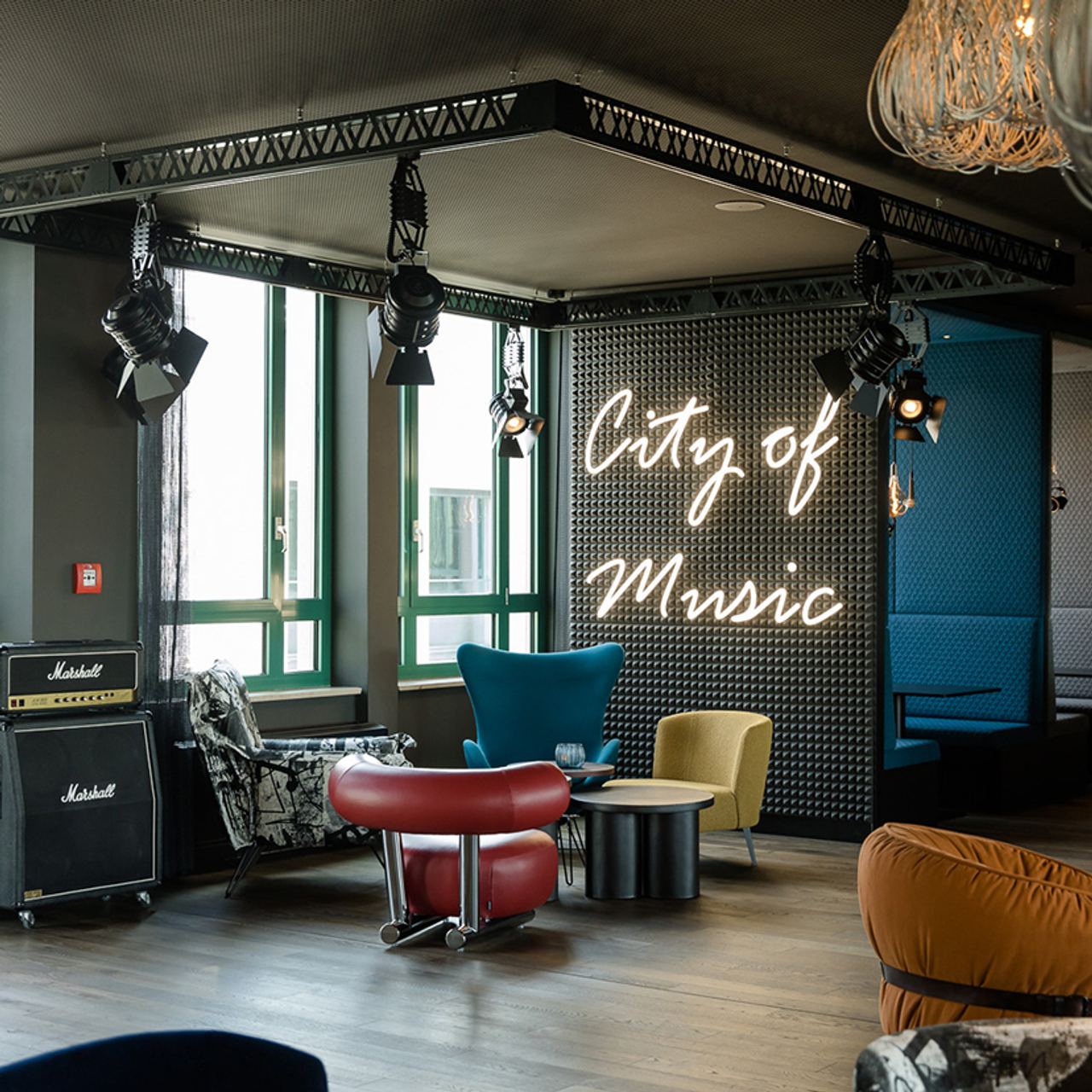 Motel One Lounge mit bequemen Sesseln, großen Fenstern und stilvoller Beleuchtung. An der Wand leuchtet in Neonschrift „City of Music“.